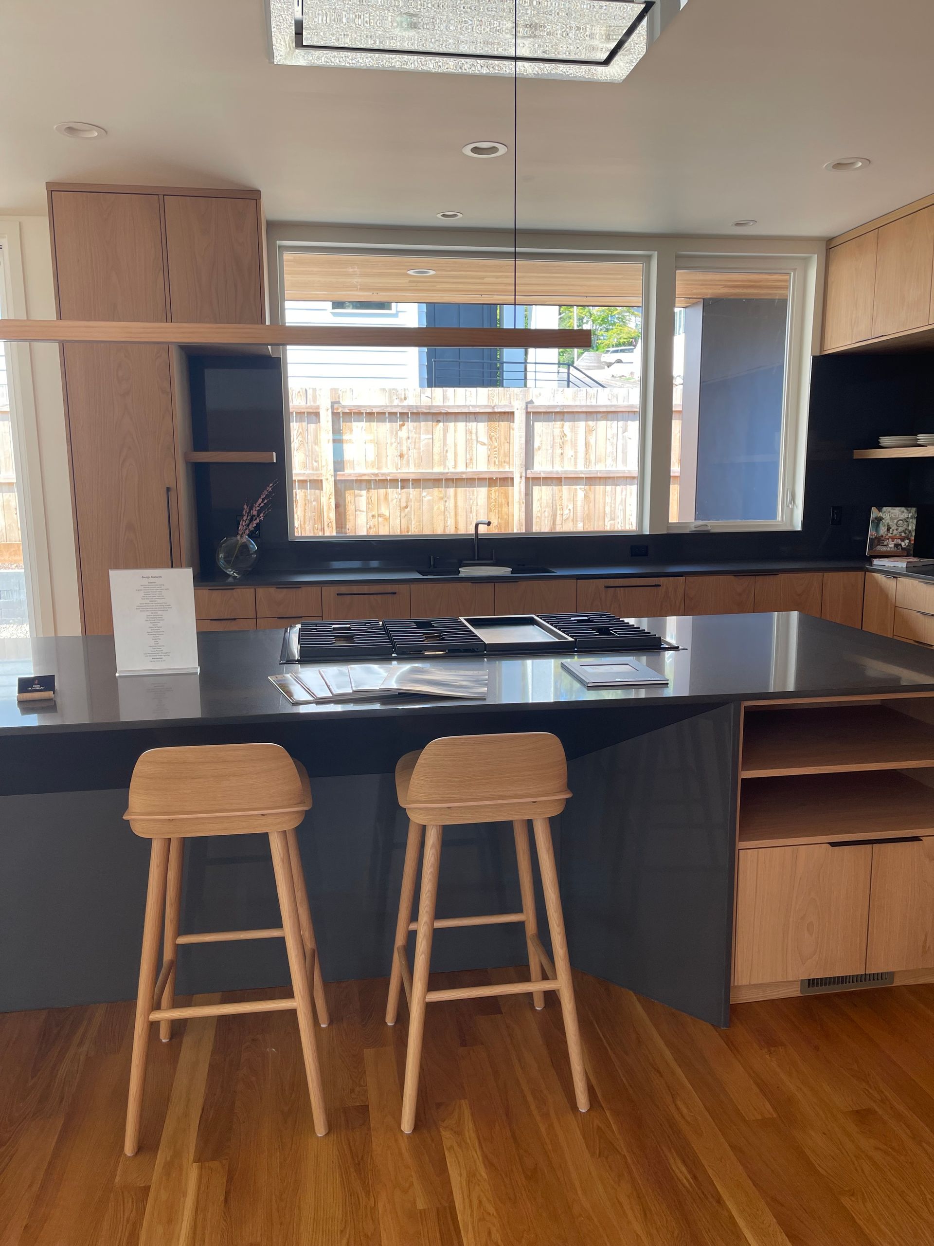 Modern kitchen with wood and dark gray cabinetry, island with two stools, and a window overlooking a backyard.