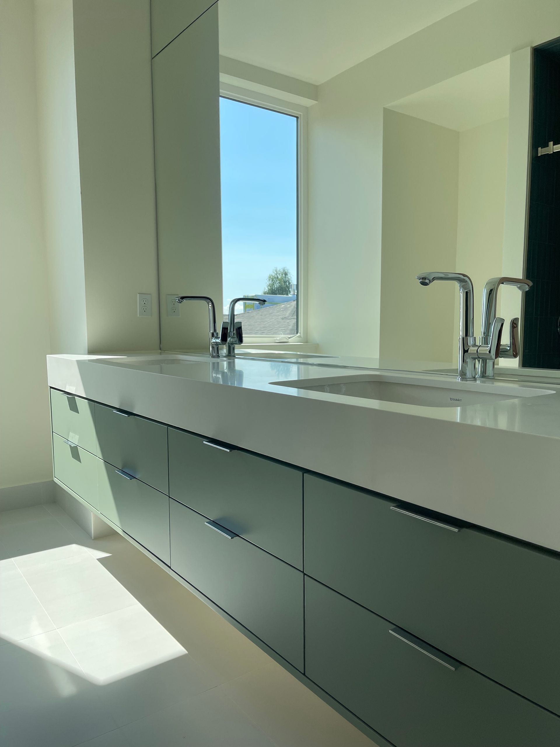 Modern bathroom with light green cabinets, white countertop, two sinks, and a window with a blue sky view.