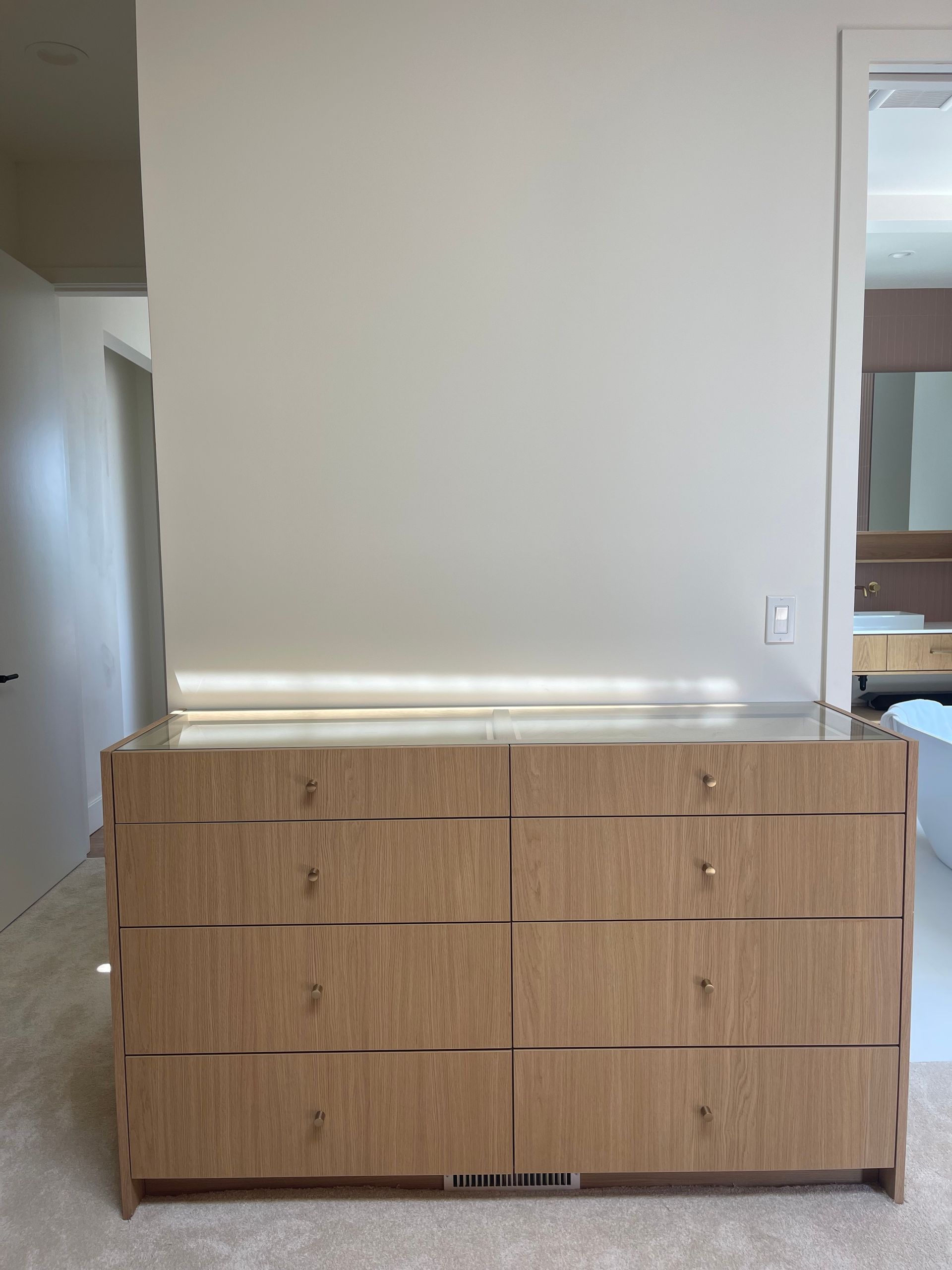 Wooden dresser against a white wall with recessed lighting. A doorway and bathroom are visible.