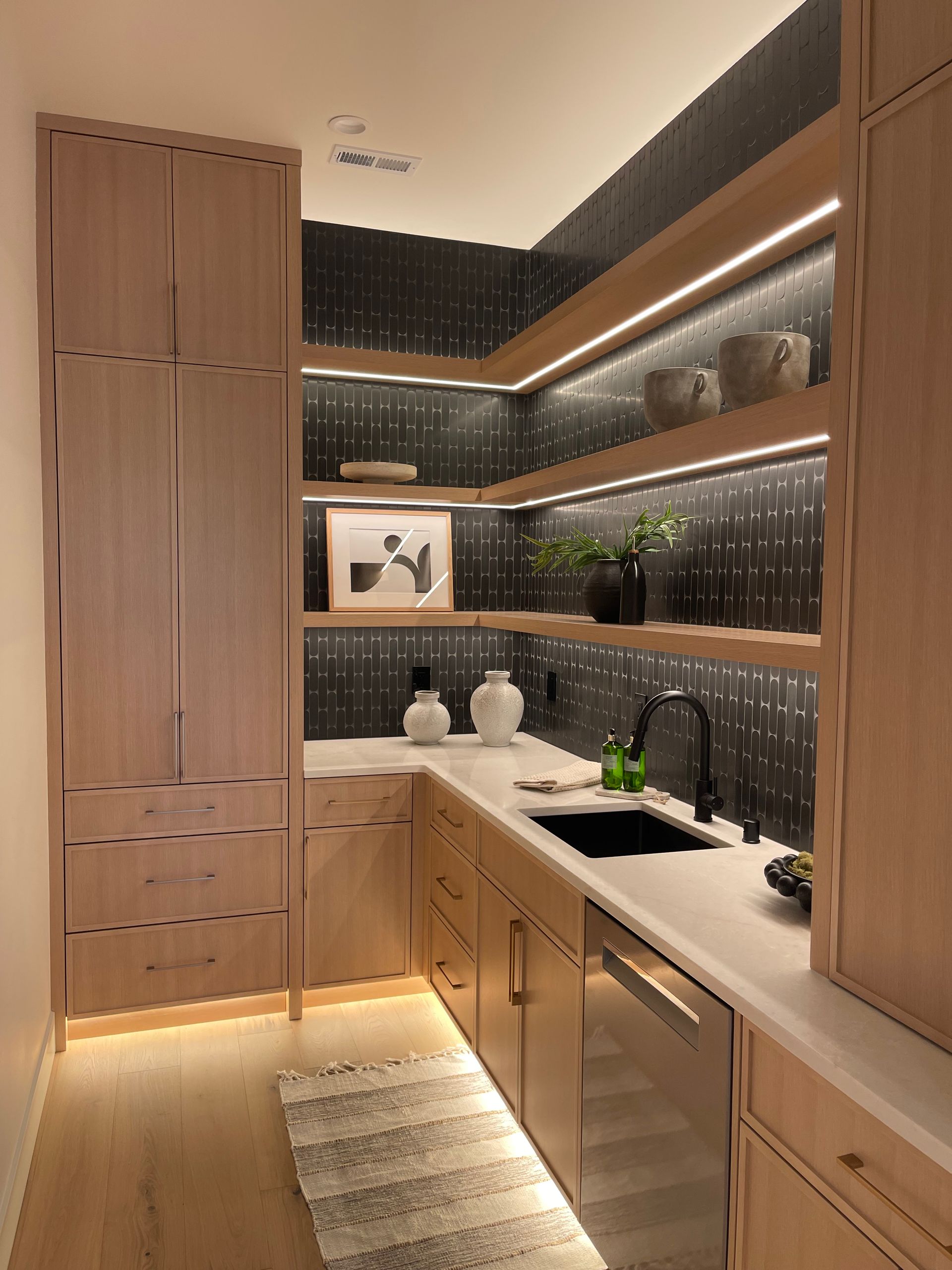 Modern kitchen with light wood cabinets, dark tiled backsplash, under-cabinet lighting, and a narrow counter.