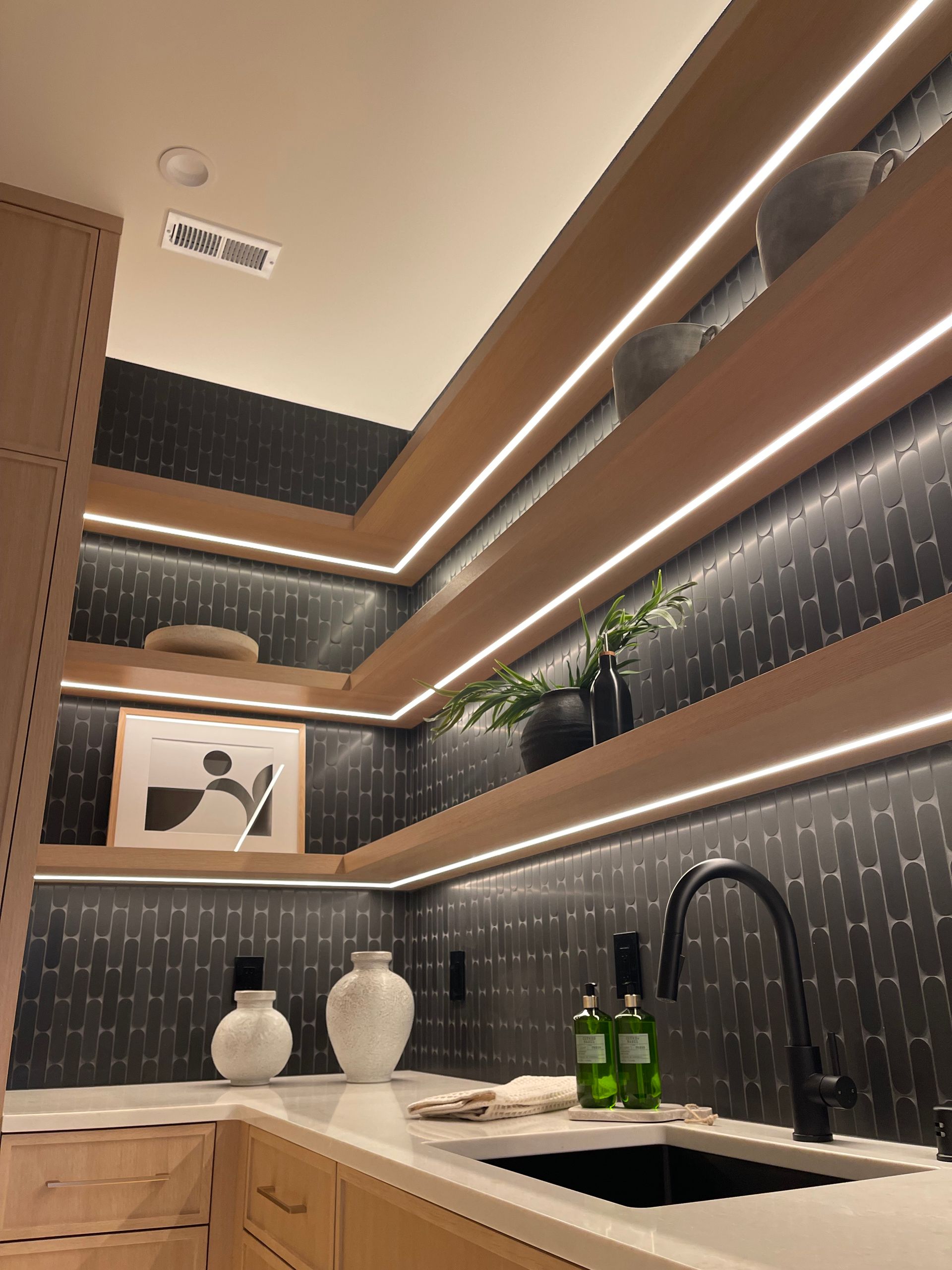 Modern kitchen corner with wood shelves, black backsplash, white vases, and sink under warm strip lighting