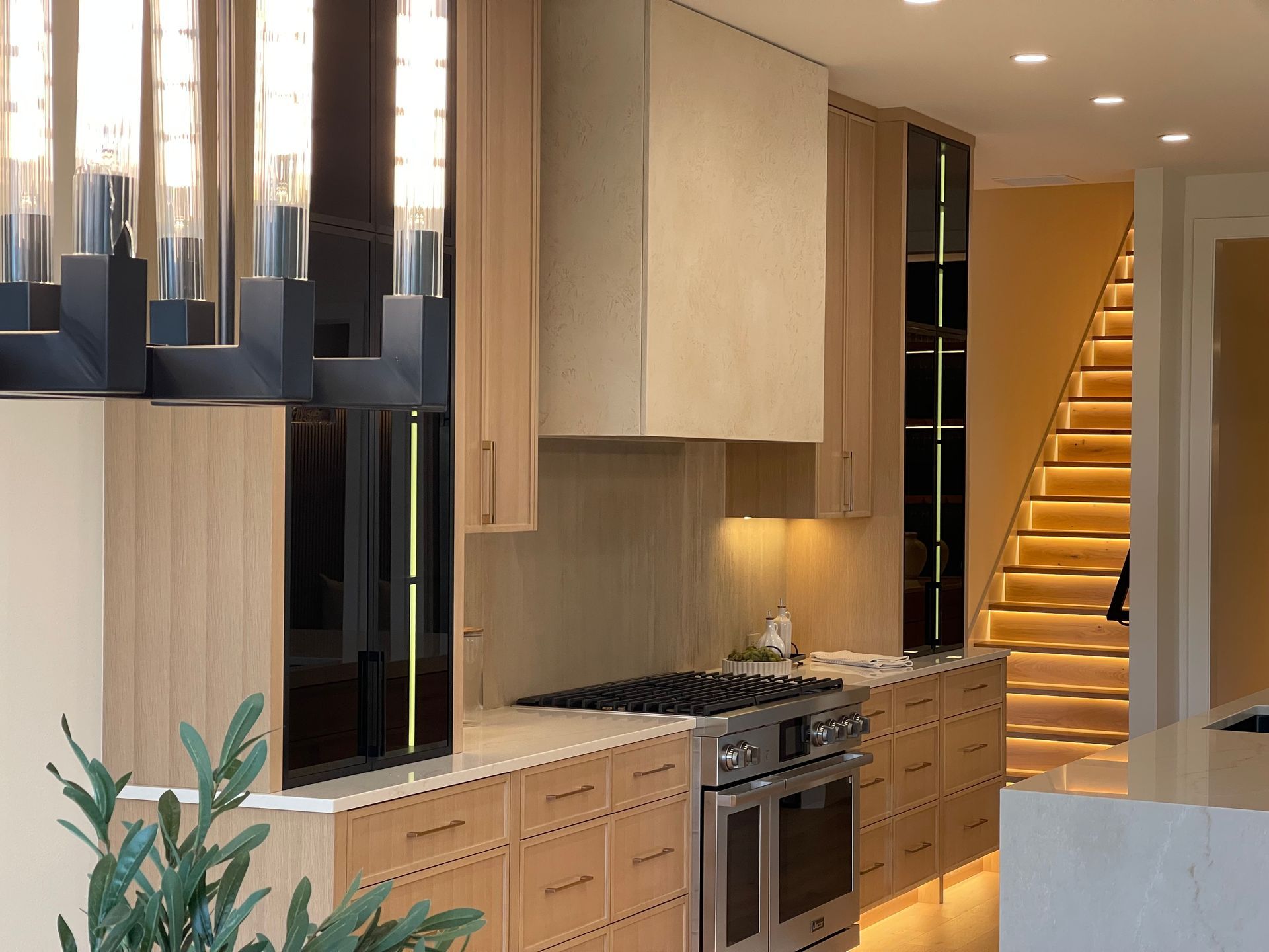 Modern kitchen with light wood cabinets, stainless stove, and staircase lit by warm lights in the background