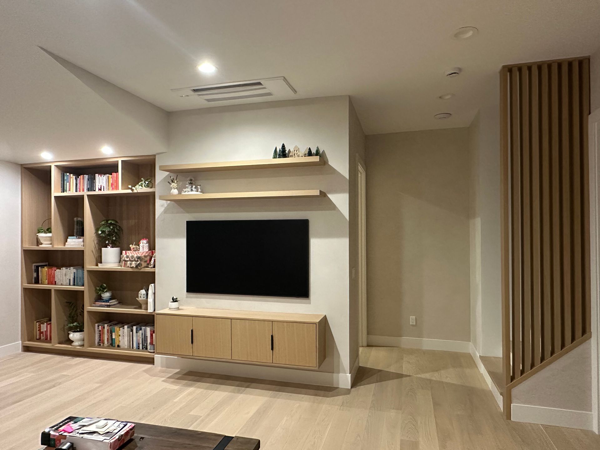 Modern living room with built-in shelving, floating TV, and wooden accents. Light wood floors and neutral walls.