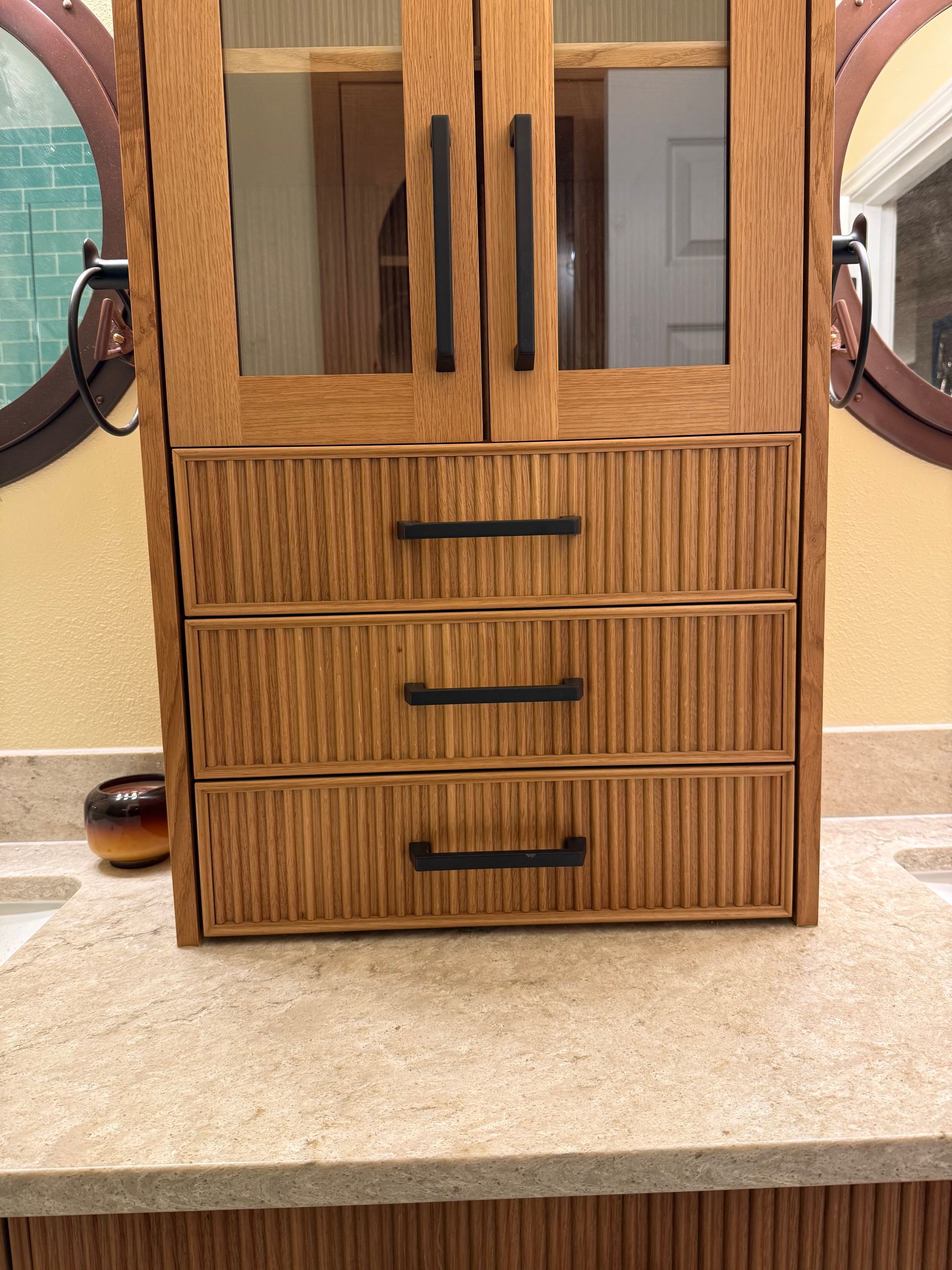 Wooden bathroom cabinet with glass doors and three drawers, set on a beige countertop