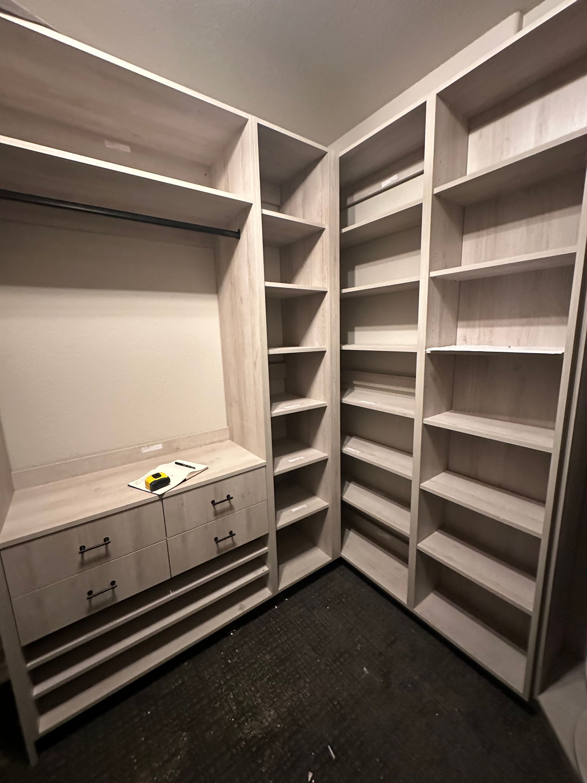 Built-in light-colored wooden closet with shelves, drawers, and a hanging rod, in a room with dark flooring.