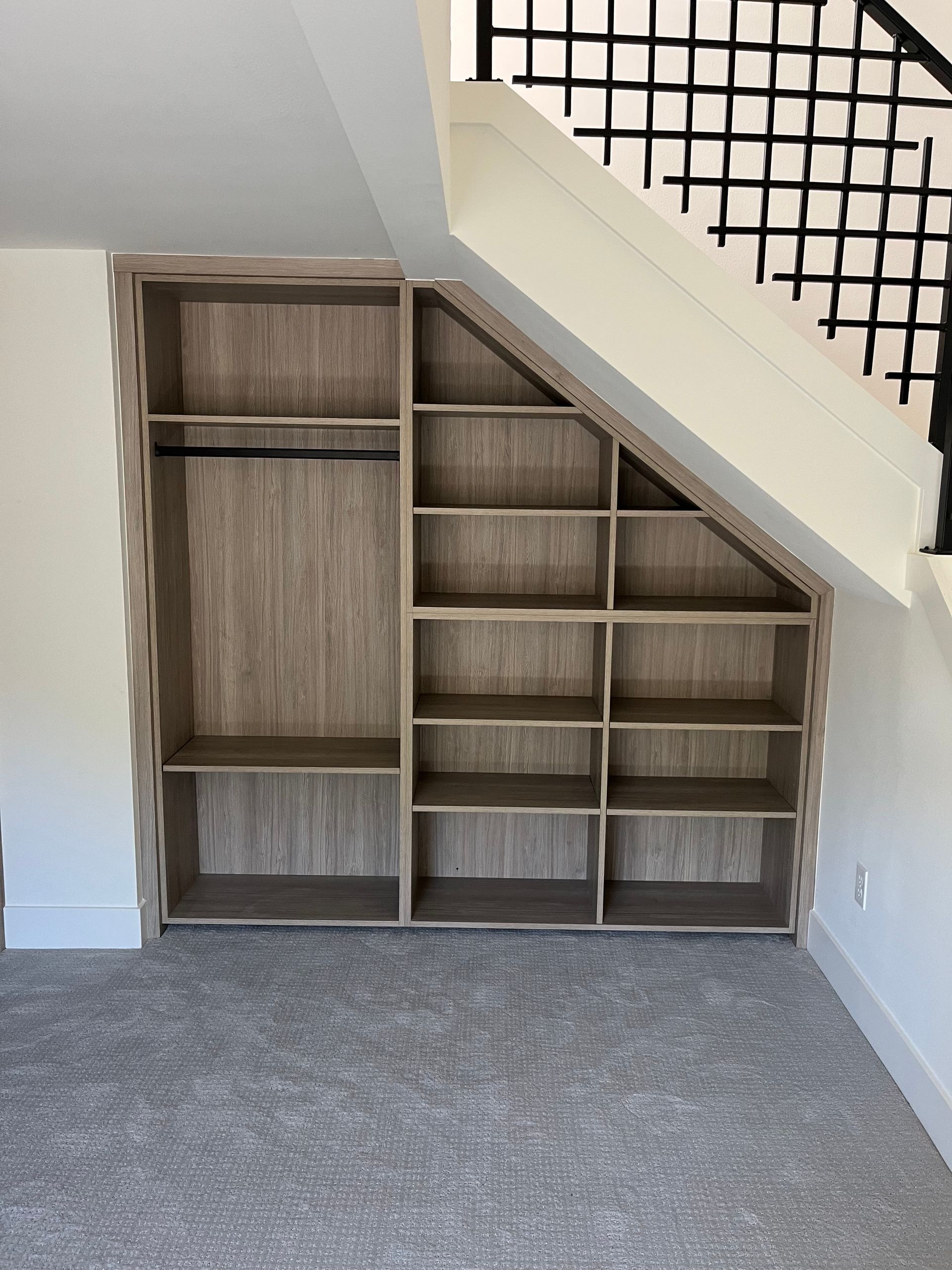 Built-in wooden shelving unit under a staircase. Shelves and hanging rod in a neutral-colored room with carpet.