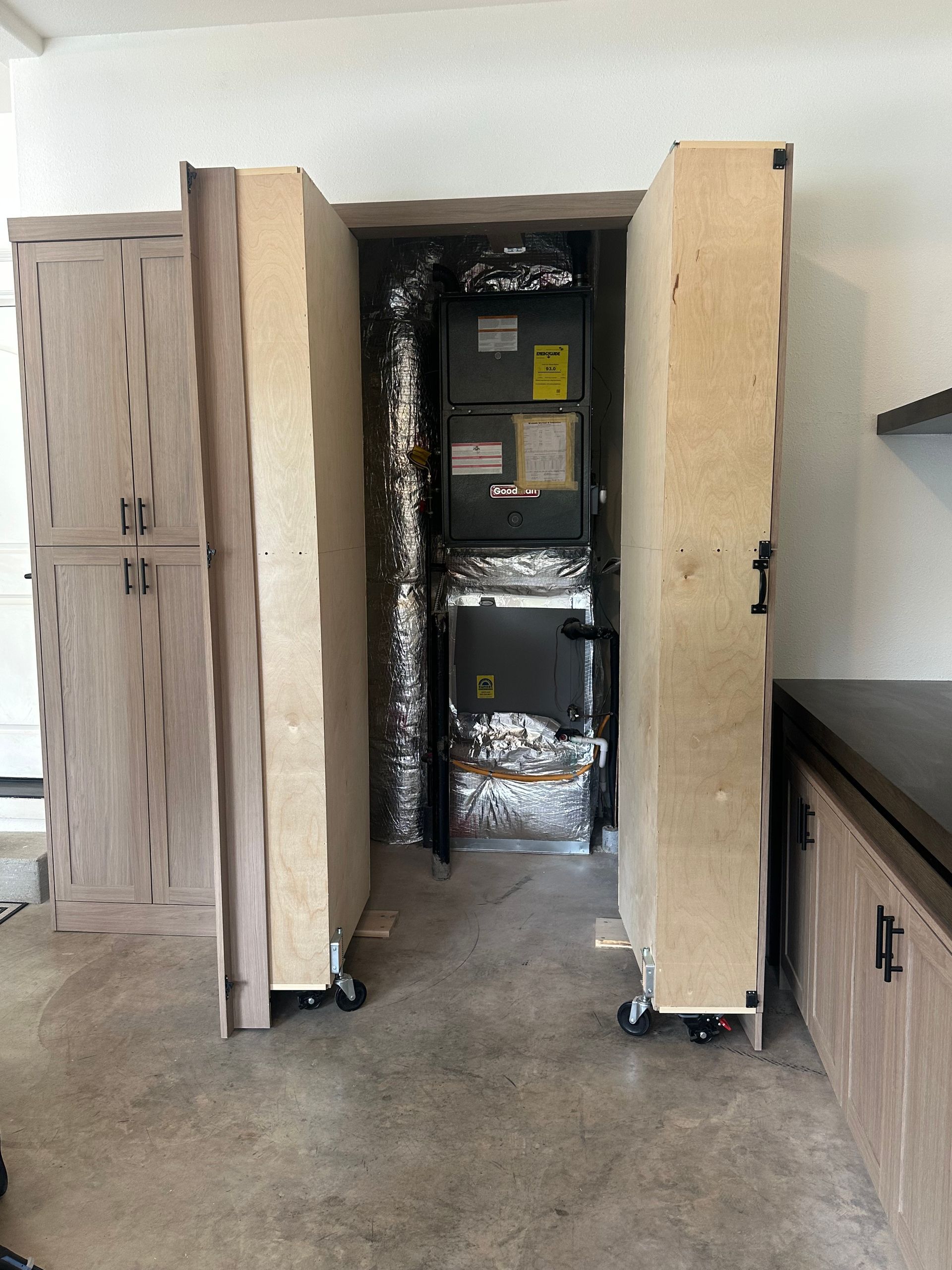 Garage with HVAC unit between two cabinet-like structures on wheels, with a cabinet on the left.