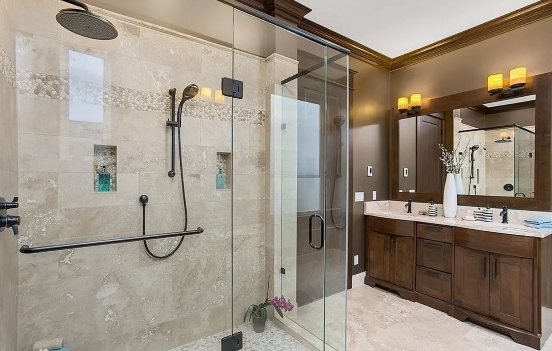 Modern bathroom with glass shower, double vanity, and brown wooden cabinets. Beige tile walls and dark fixtures.