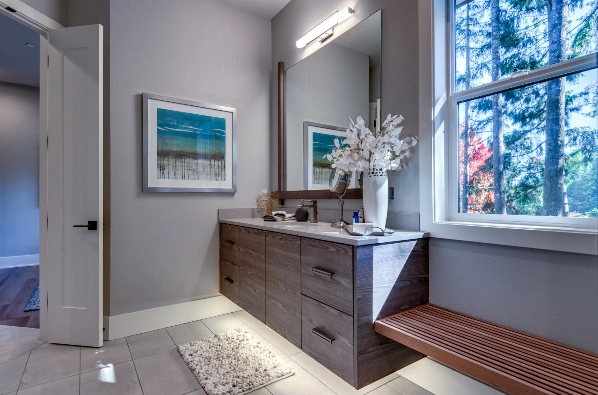 Modern bathroom with gray walls, floating vanity, large mirror, and a window overlooking trees.
