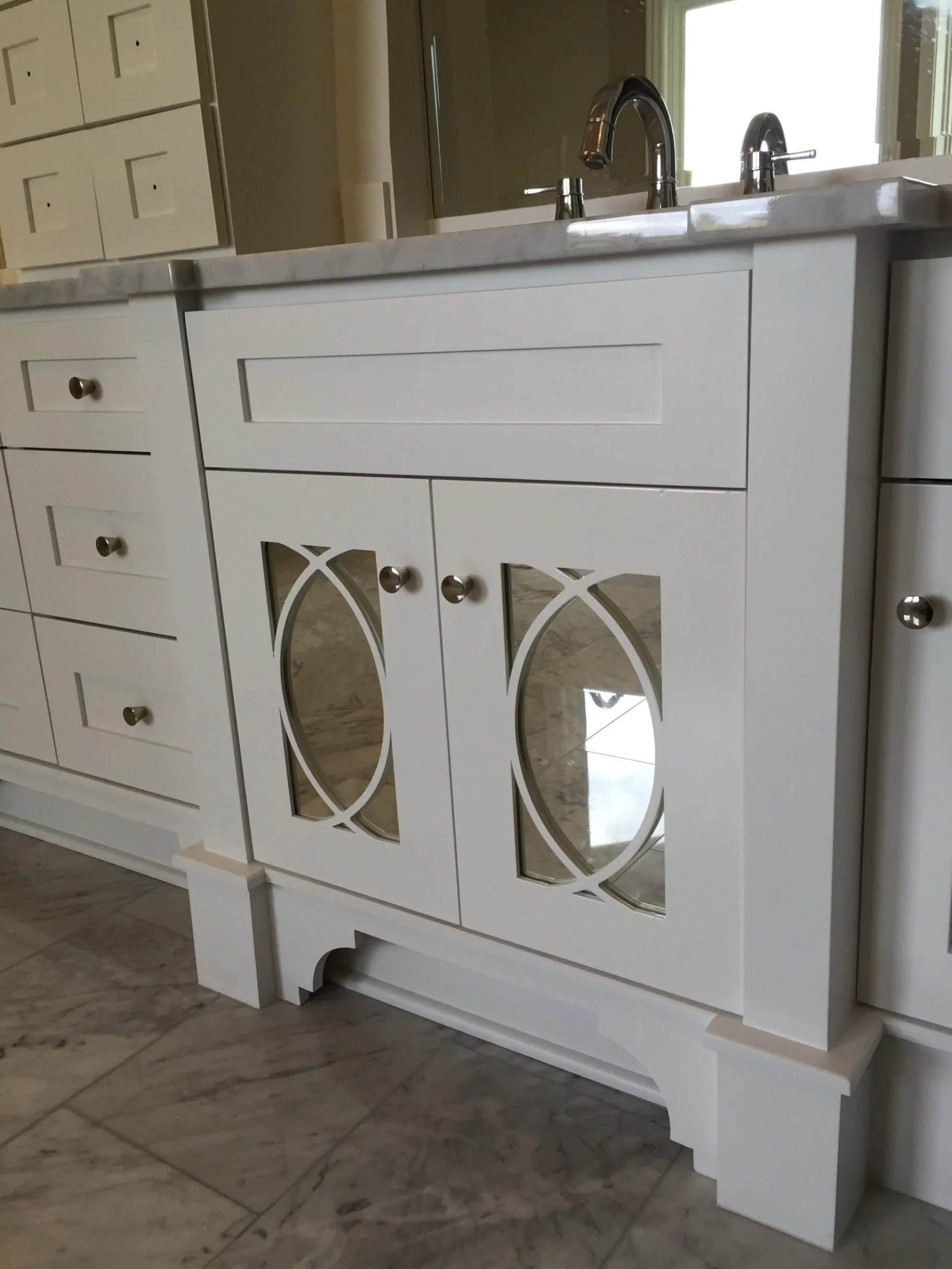 White bathroom vanity with oval mirrored cabinet doors. Marble countertop, silver faucet.