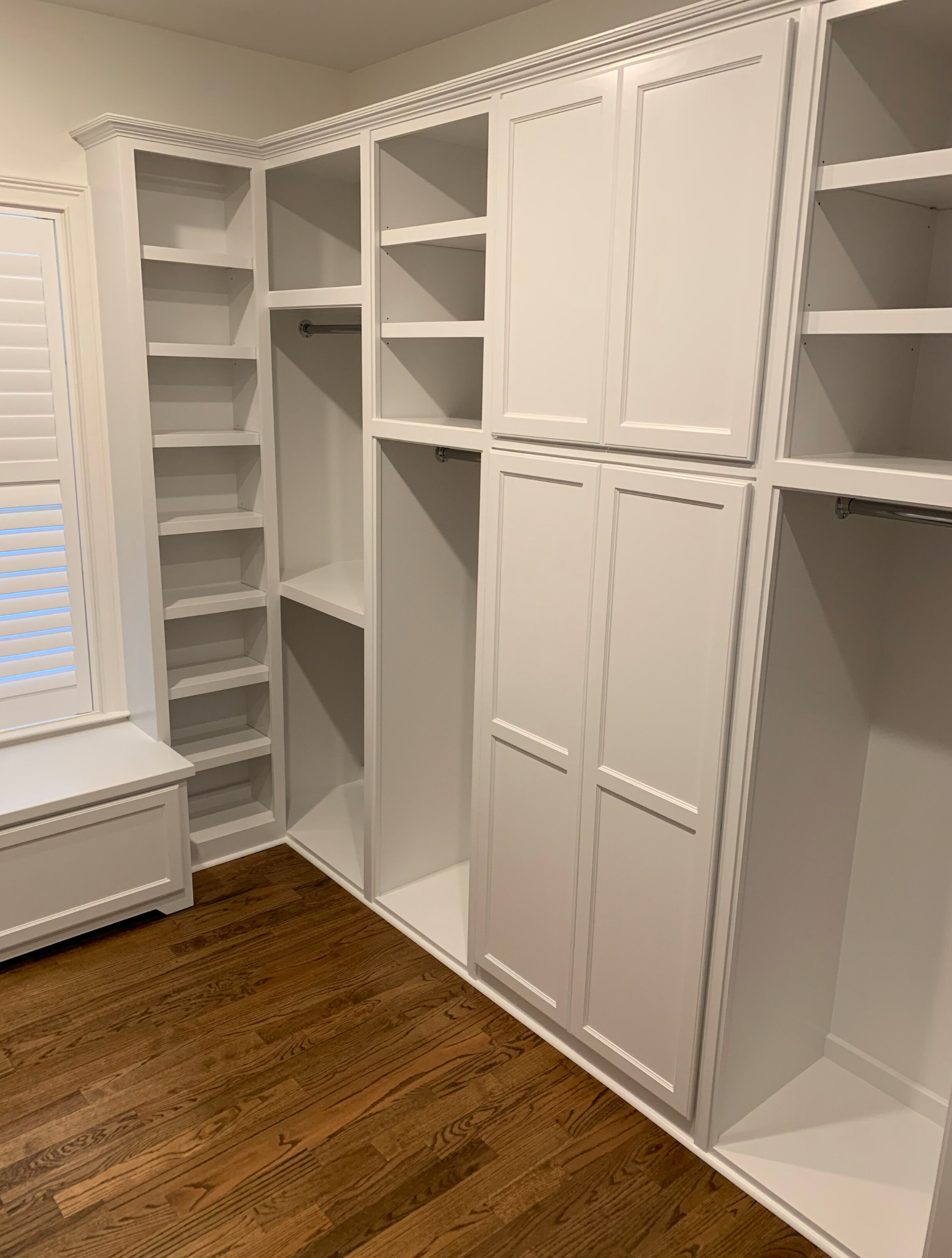 White built-in closet with shelves, cabinets, and hanging rods on a hardwood floor. A window with shutters is on the left.