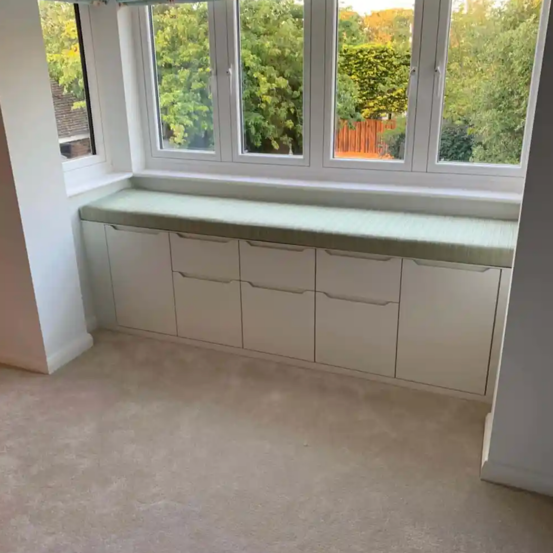 Built-in white storage cabinets below a window with a light green countertop; room with light-colored carpet.