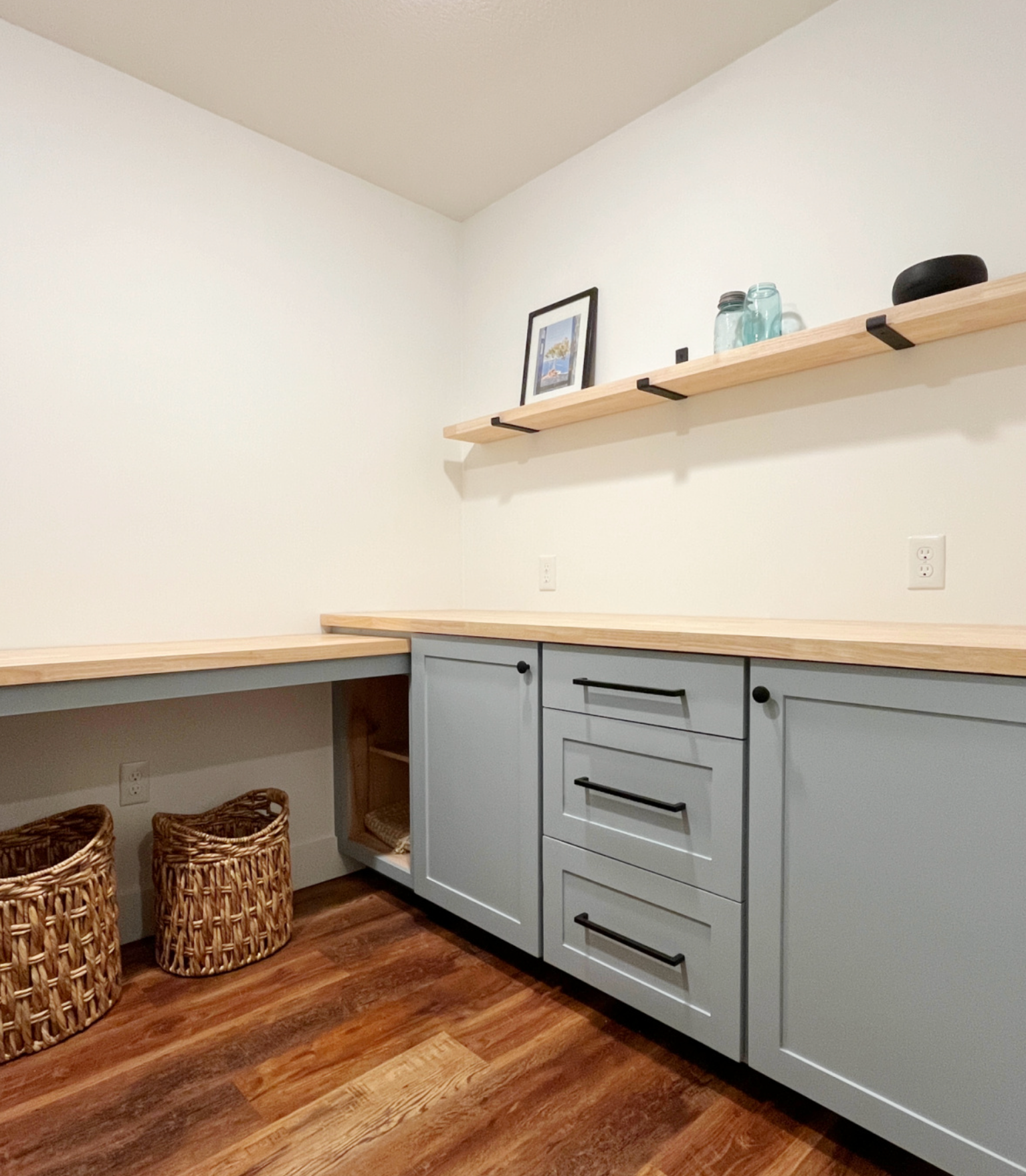 Gray cabinets with a wood countertop, floating shelf, and two wicker baskets in a home office.