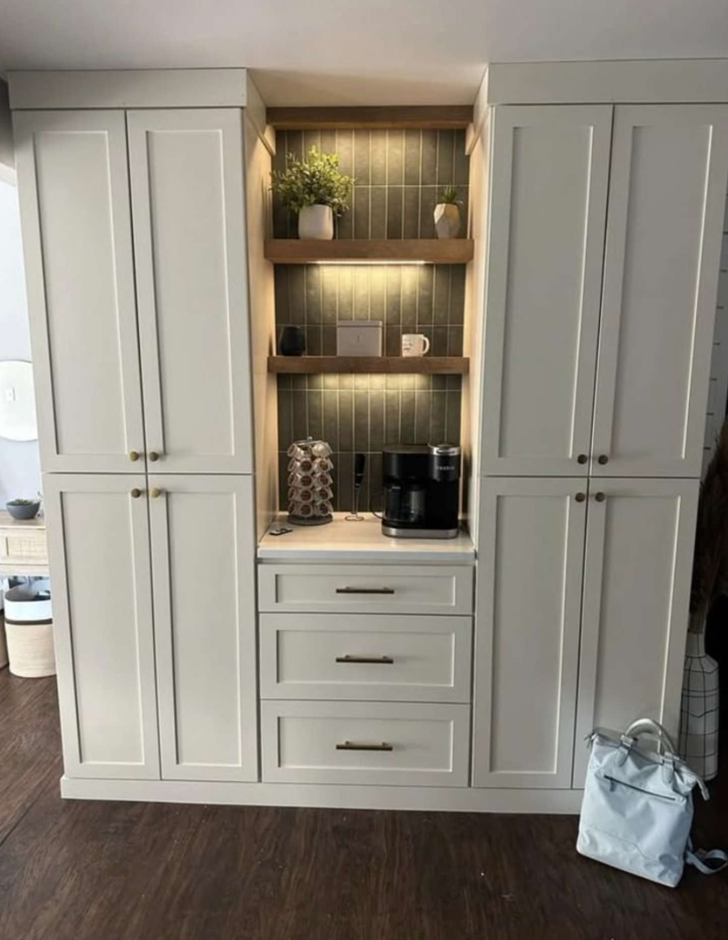 Cream-colored built-in cabinet with coffee station. Drawers, shelves, coffee maker, plants. Wooden shelves with green tile.