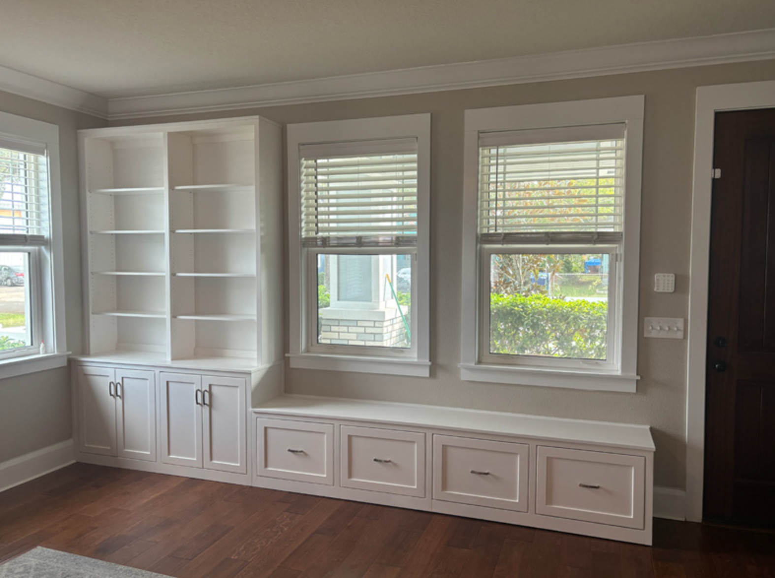 Built-in white cabinets with bookshelves, drawers, and window seat under two windows. Dark wood floor.