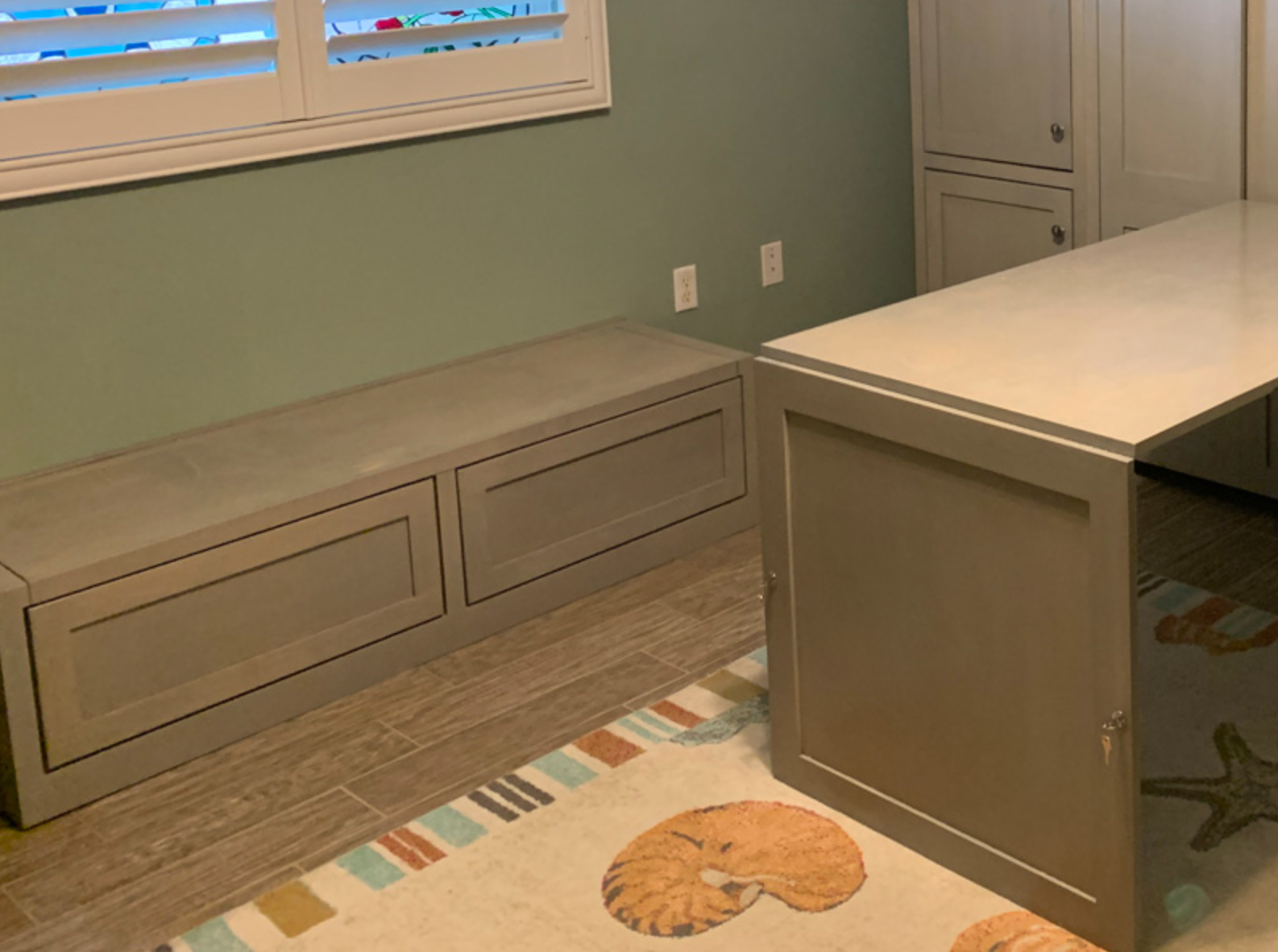 Built-in storage bench and desk in a room with a window and a rug. The walls are green and the furniture is gray.