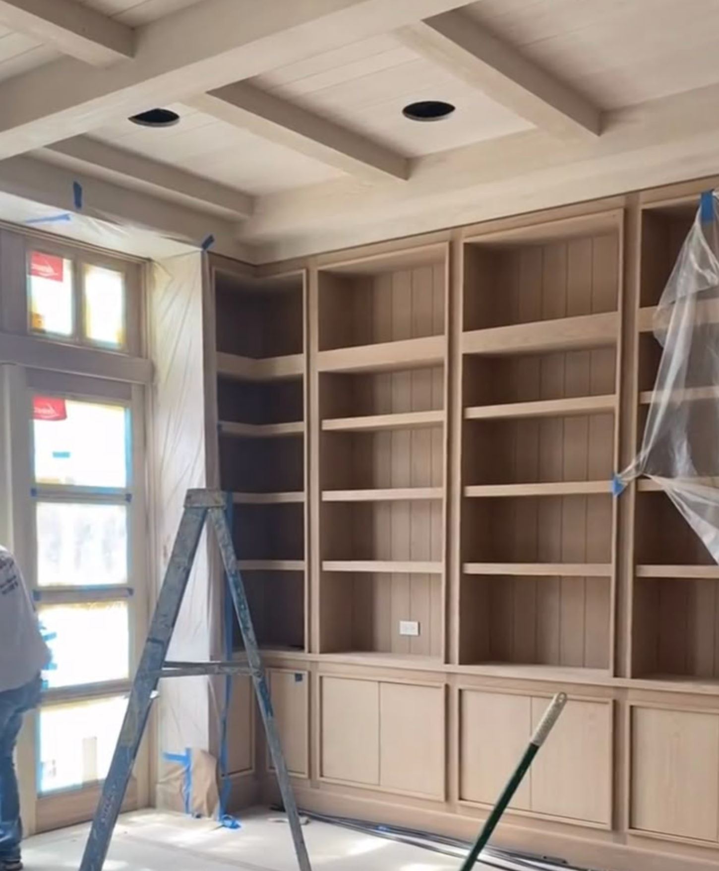 Built-in wood shelving and cabinets in a room under renovation, with ladders and plastic sheeting.
