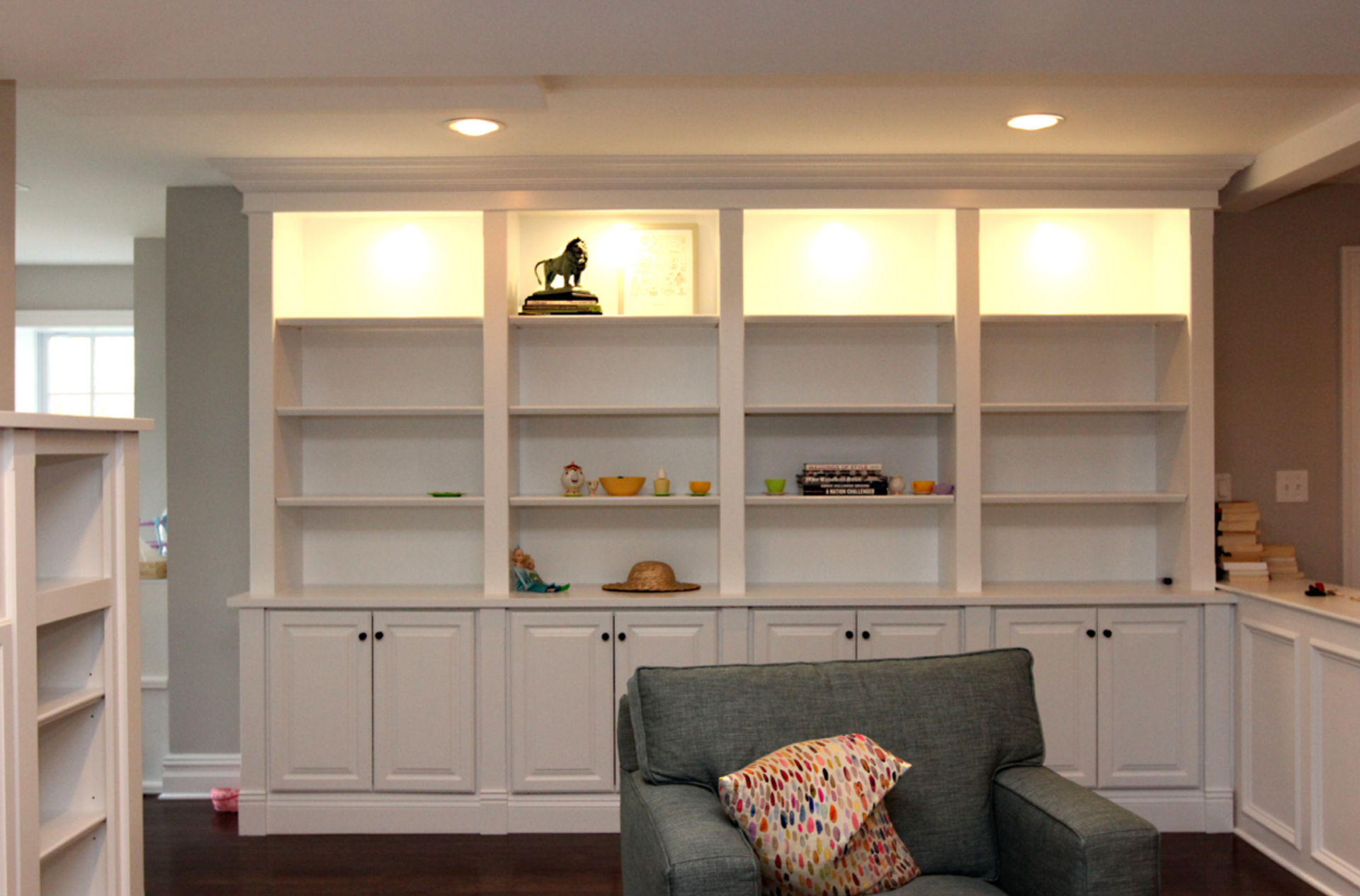 Built-in white shelving and cabinets in a living room, with a green chair in front and recessed lights above