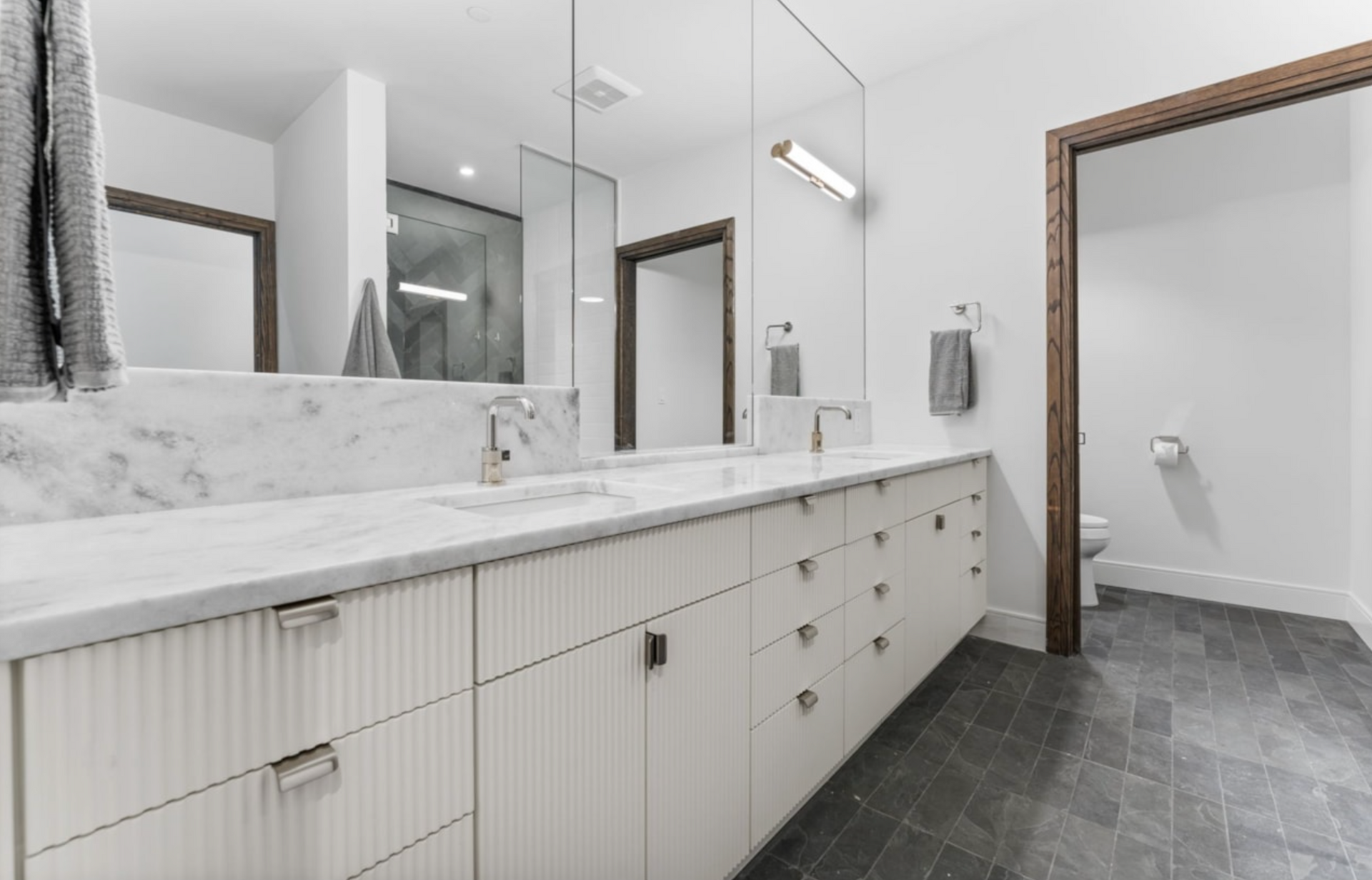 Modern bathroom with white cabinets, marble countertop, large mirrors, and dark gray tile floor.