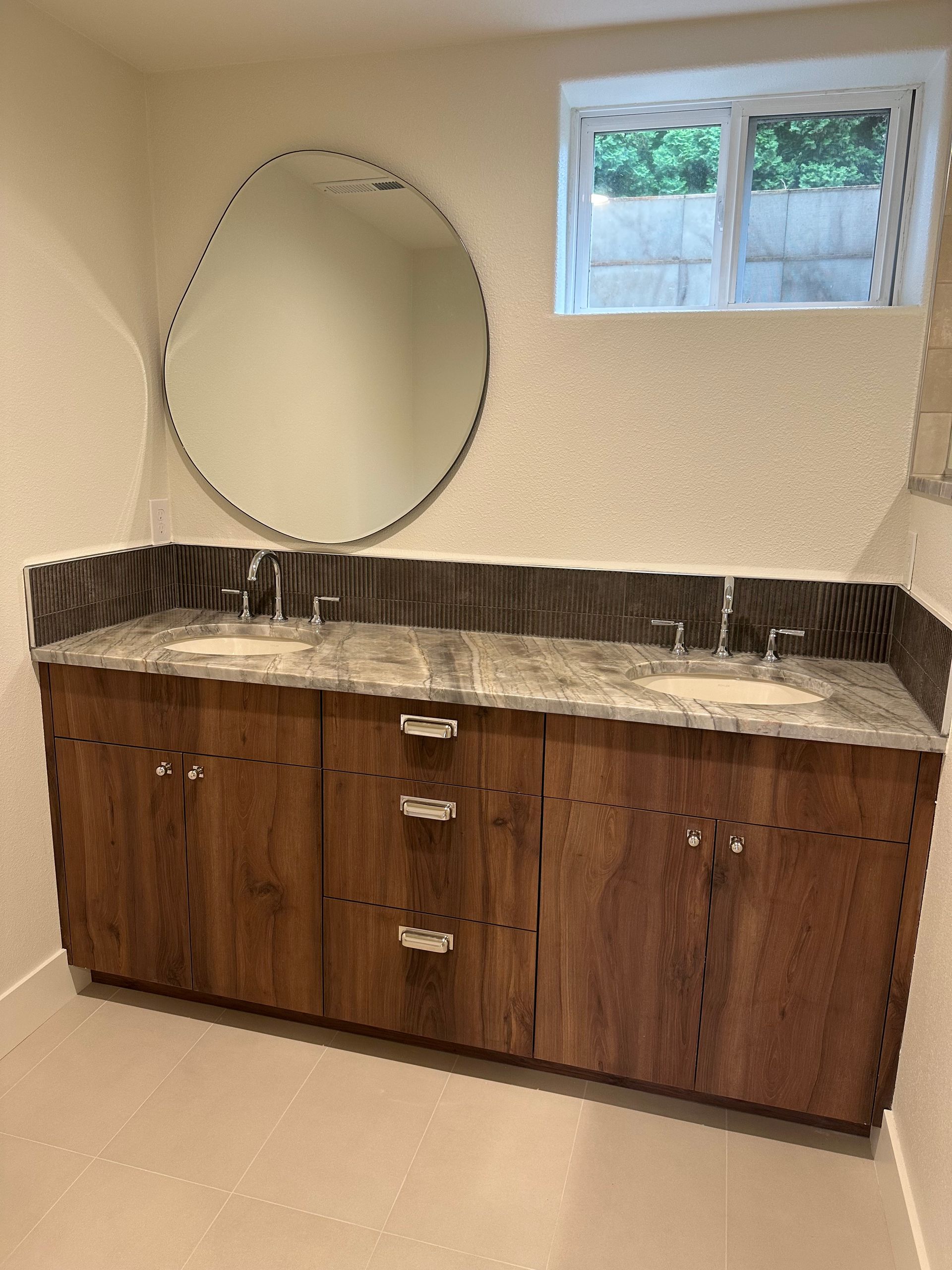 Bathroom with wooden double vanity, round mirror, and small window.