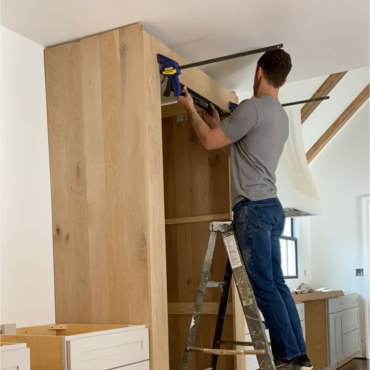 Man on ladder using clamps to attach wood to cabinet in kitchen.