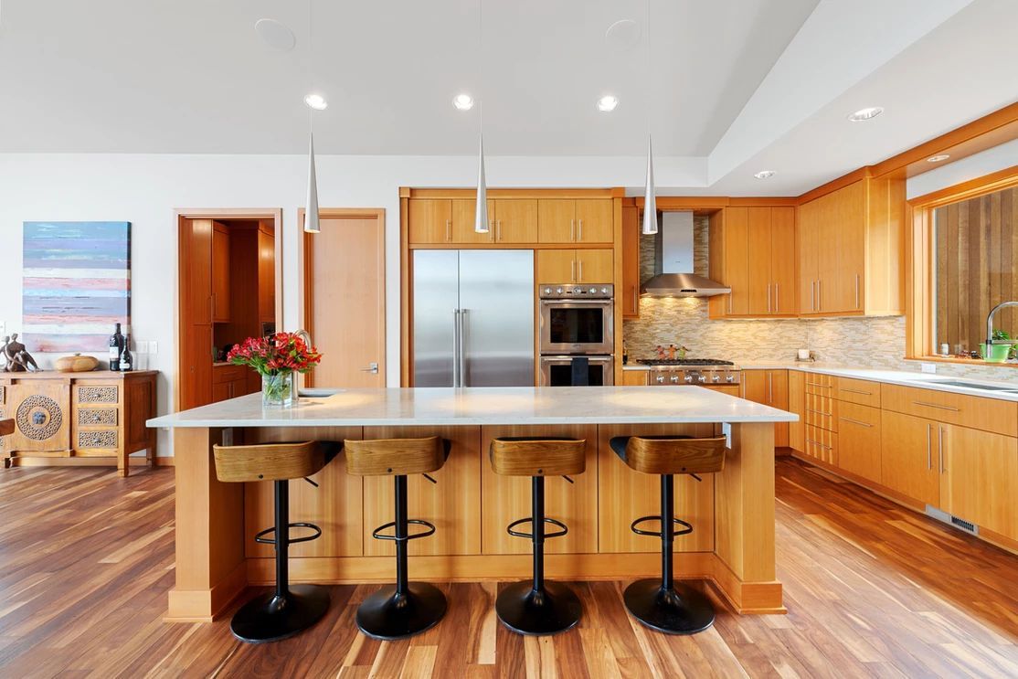 Kitchen with light wood cabinets, island with stools, stainless steel appliances, and wood floor.