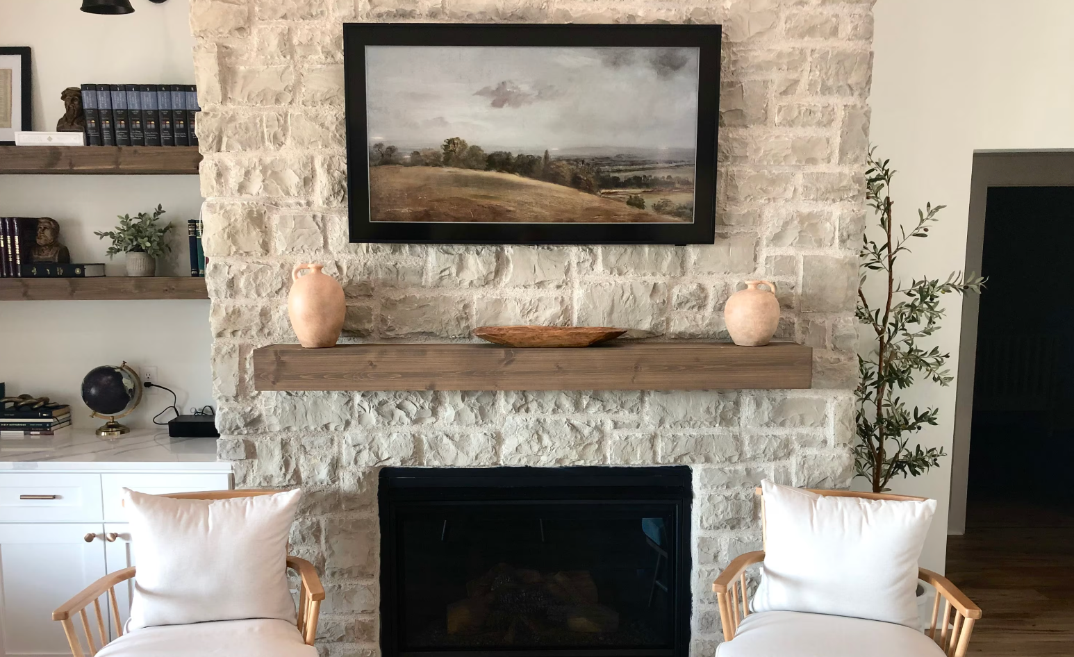 Fireplace with stone facade, painting, wooden mantle, shelves, and chairs.