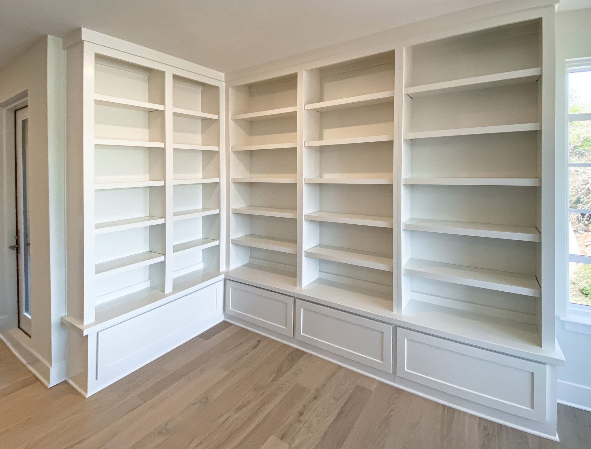 White built-in bookshelves with lower storage cabinets against a wall and a window.
