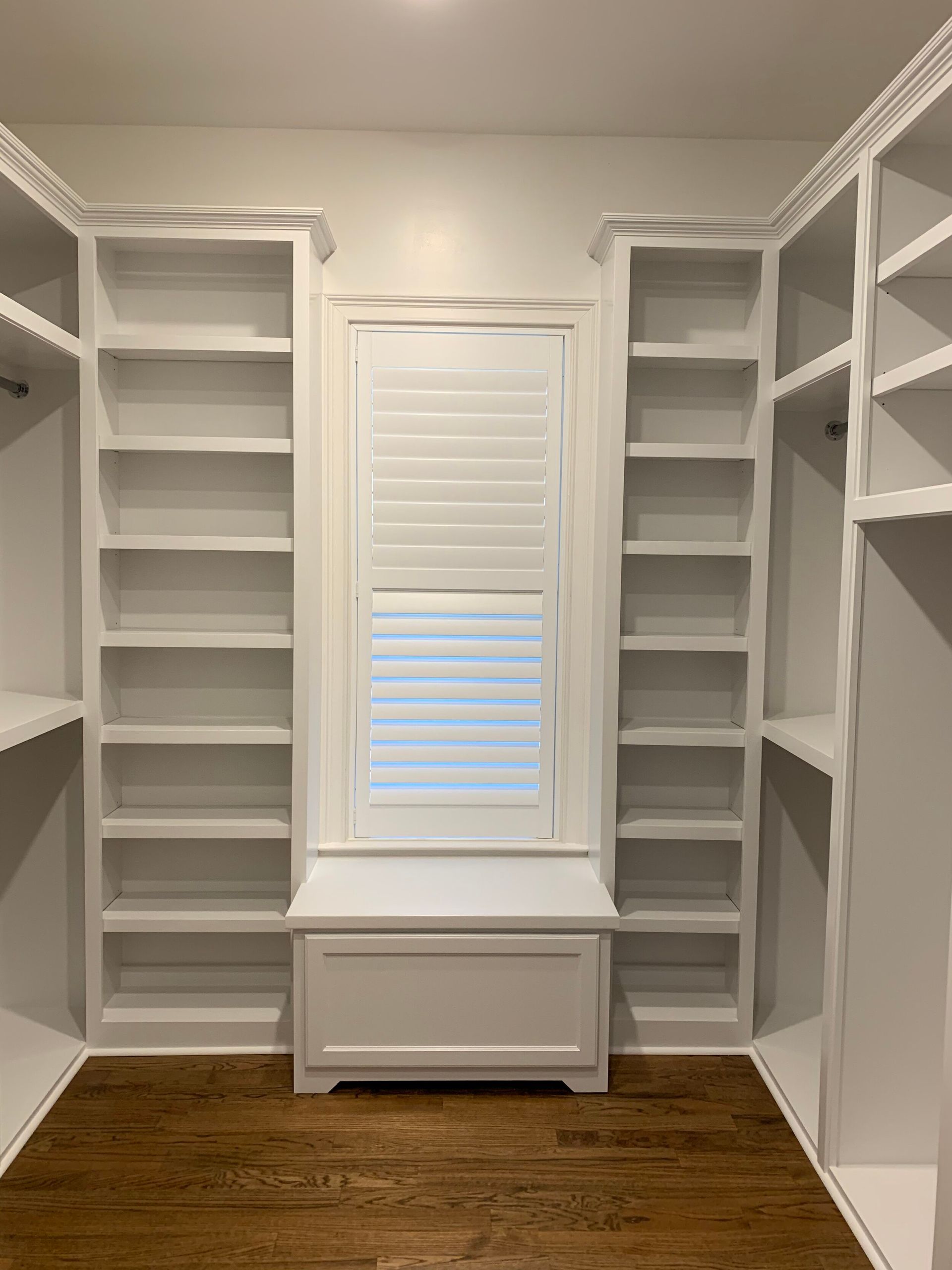 White closet with shelving on either side of a window with shutters and bench. Wood floor.