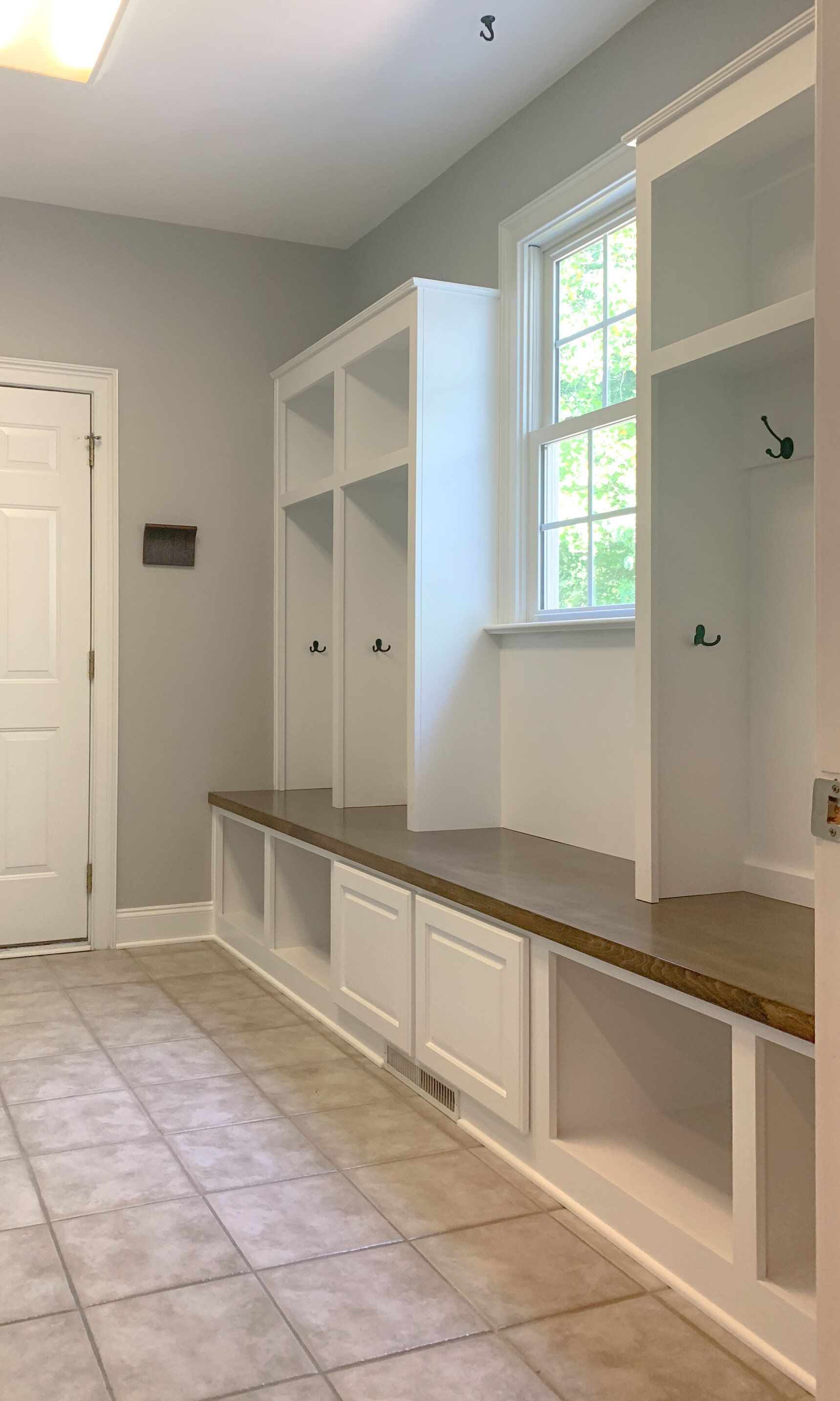A mudroom with built-in white storage, bench seating, and a window. Beige tile floor and light gray walls.