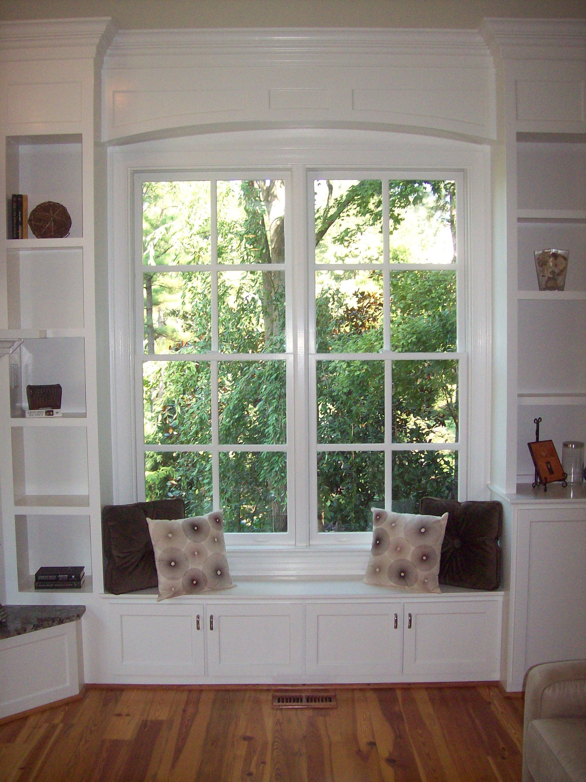 White window seat with built-in shelves. Two pillows sit on the seat. Large window with green trees.