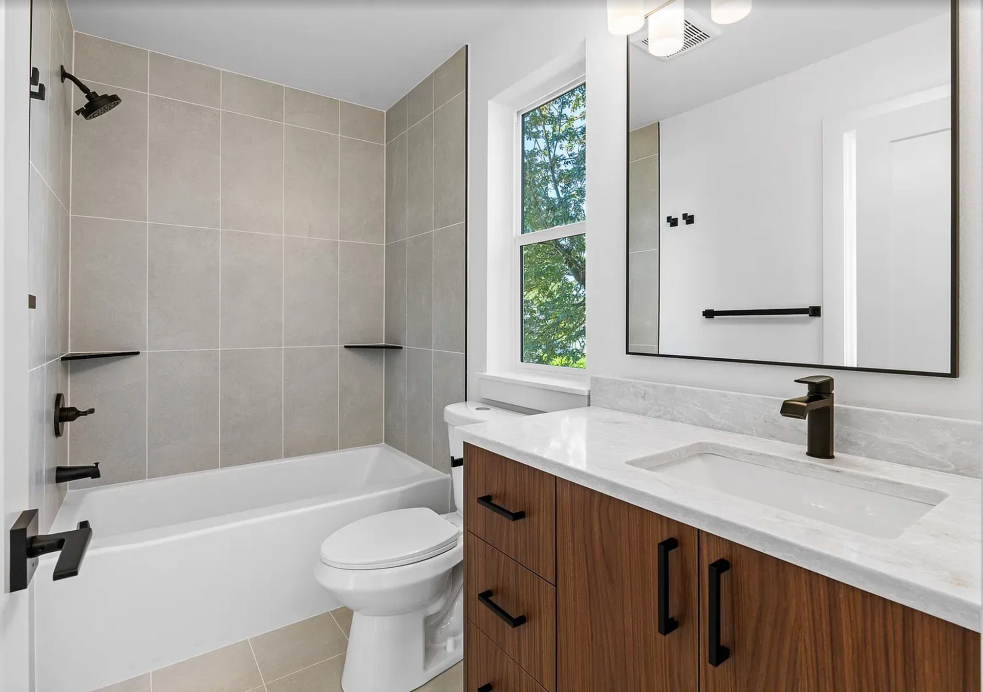 Bathroom with gray tiled walls, white tub, wooden vanity, and large mirror.