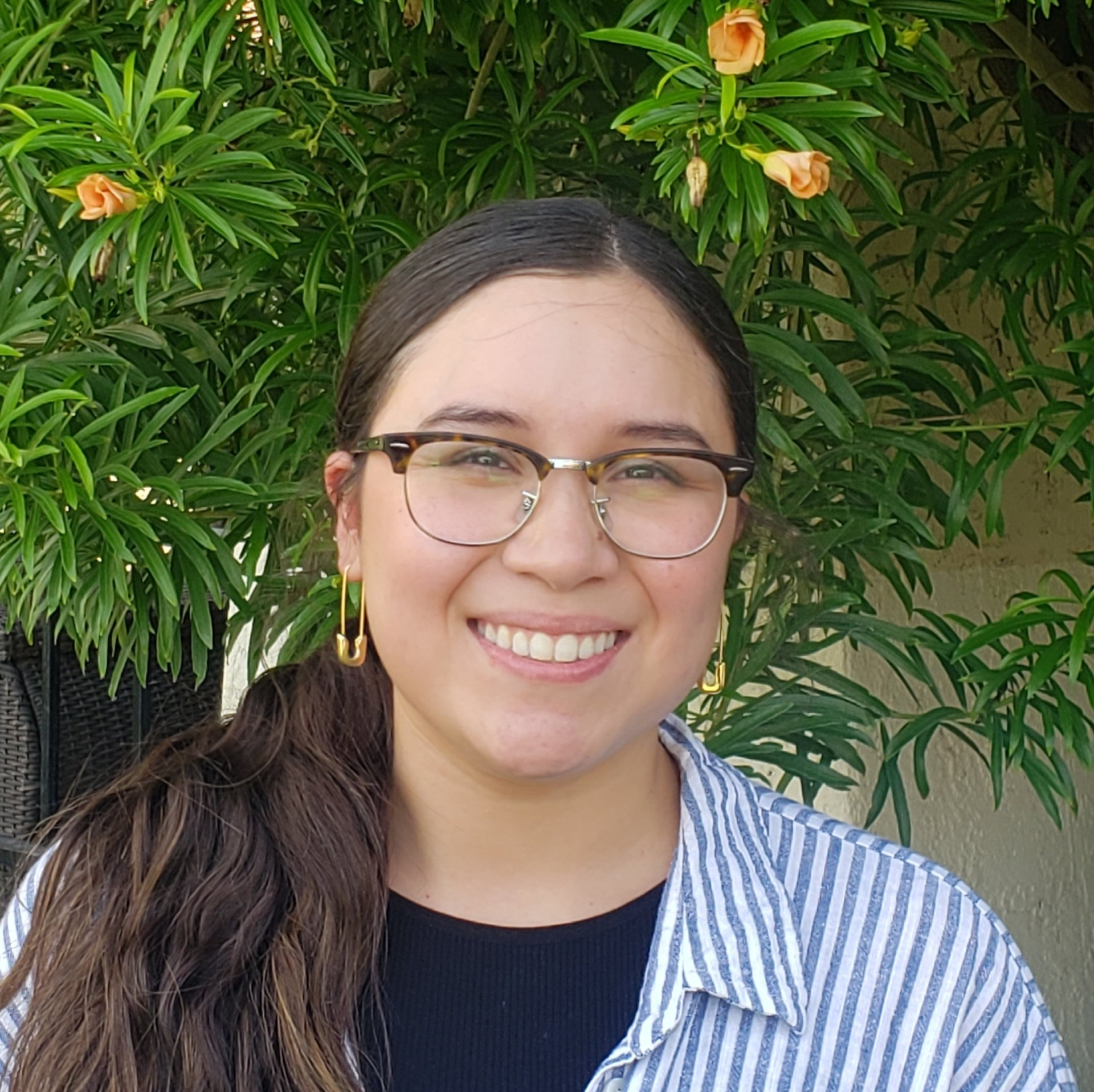 A woman wearing glasses and earrings is smiling in front of a tree.