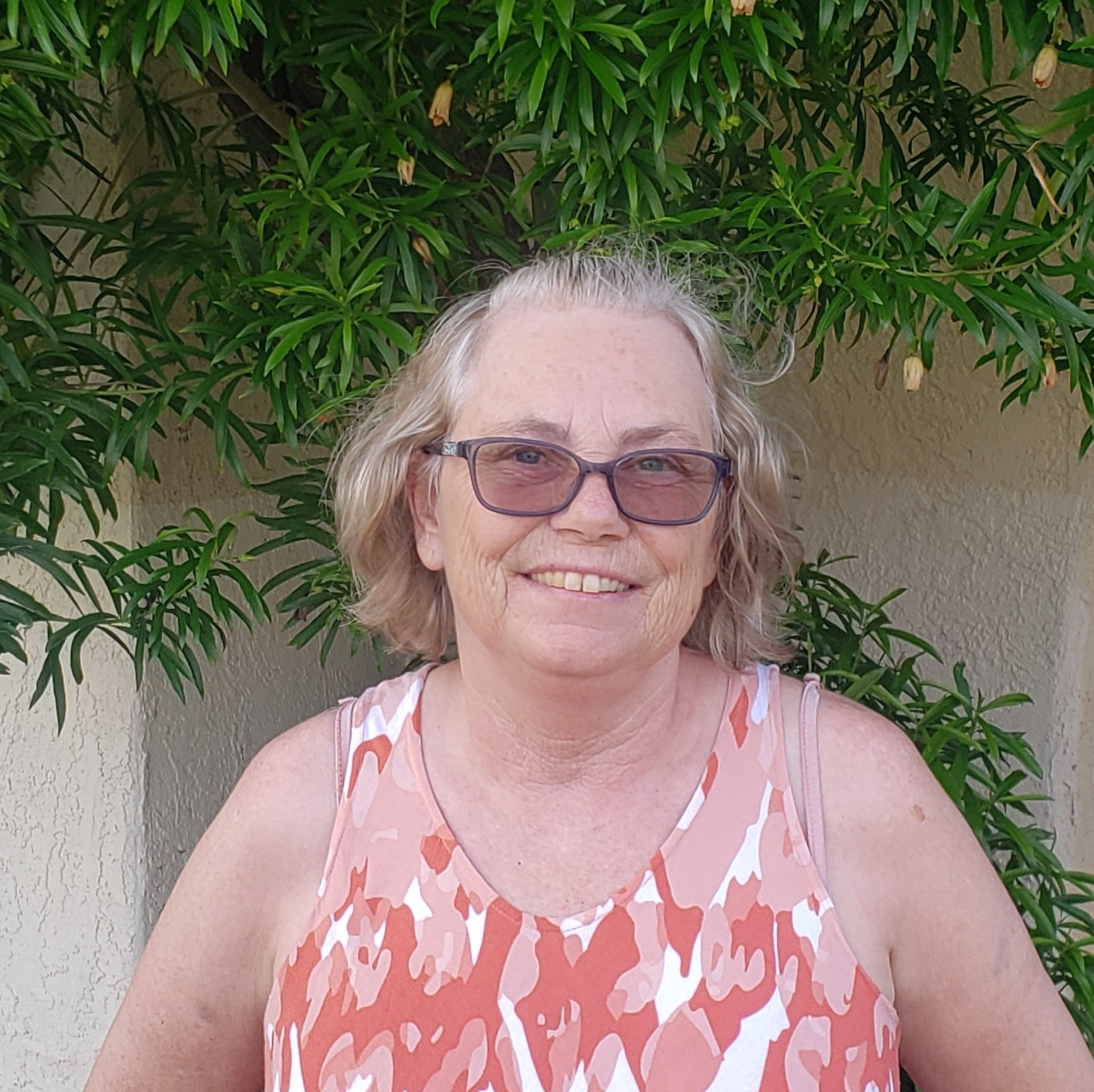 A woman wearing glasses and a tank top is smiling in front of a tree.