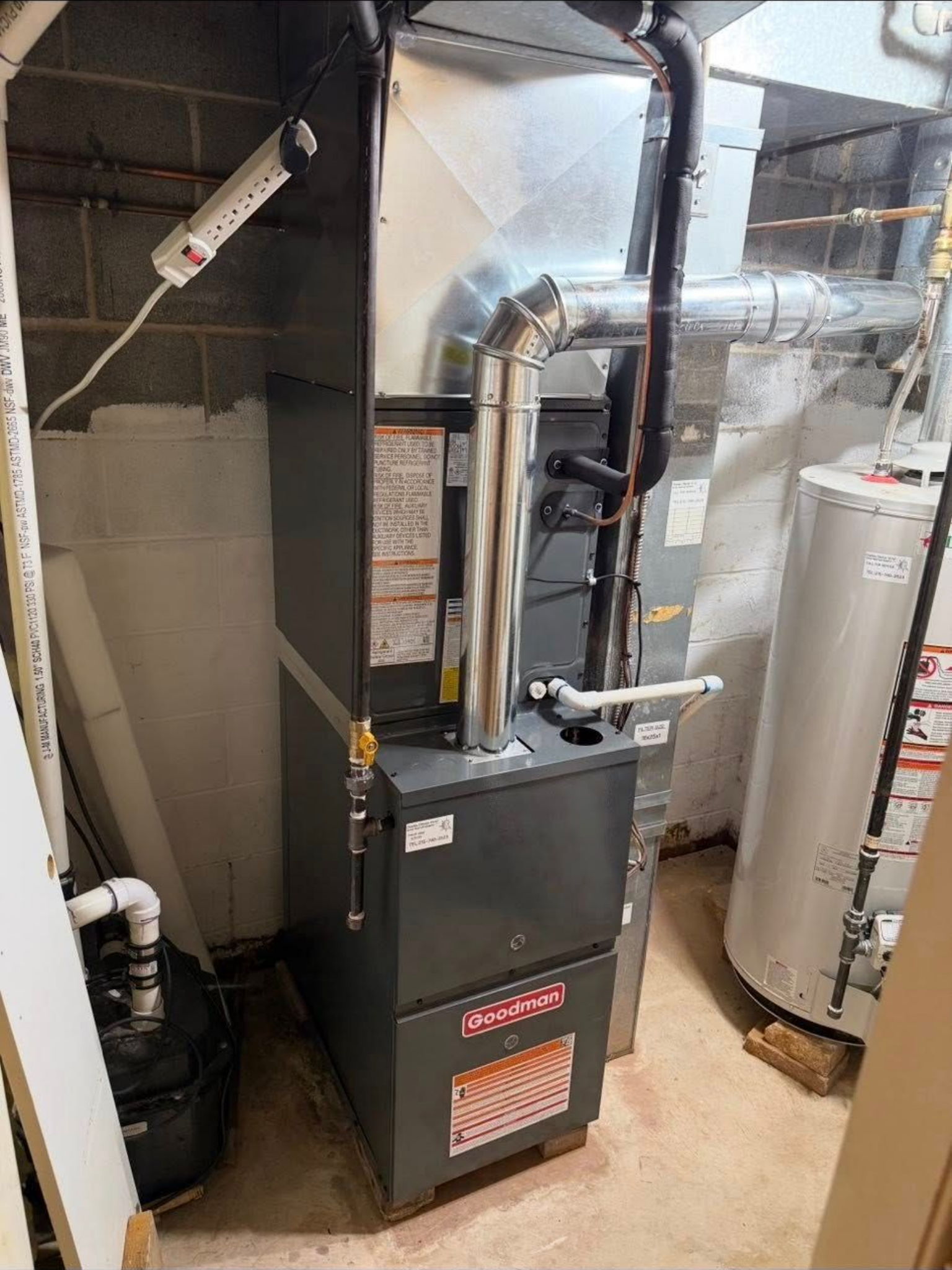 A grey residential furnace stands in a basement next to a white water heater and a black sump pump.