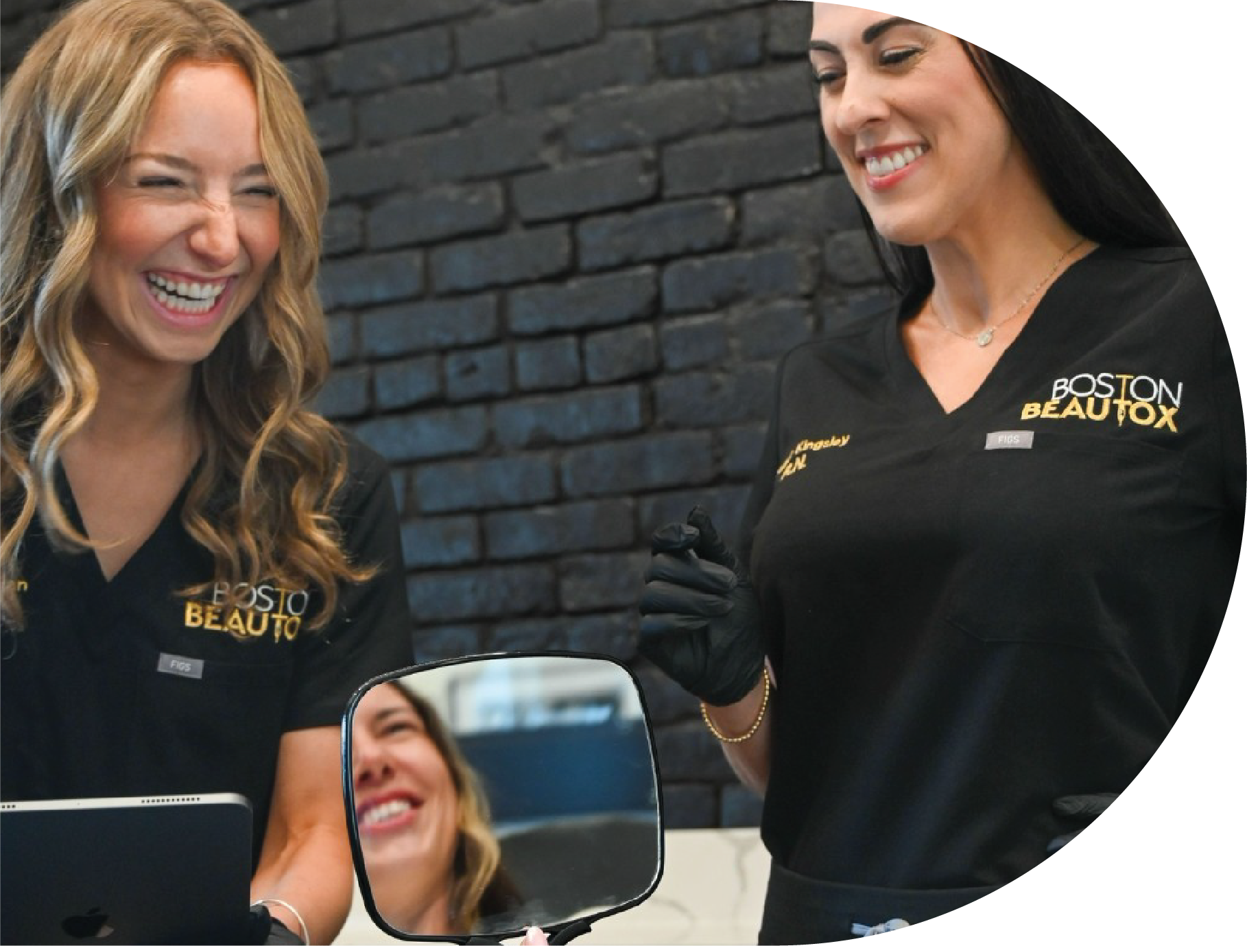 Two women in black scrubs, one holding a mirror, laughing in front of a dark brick wall.