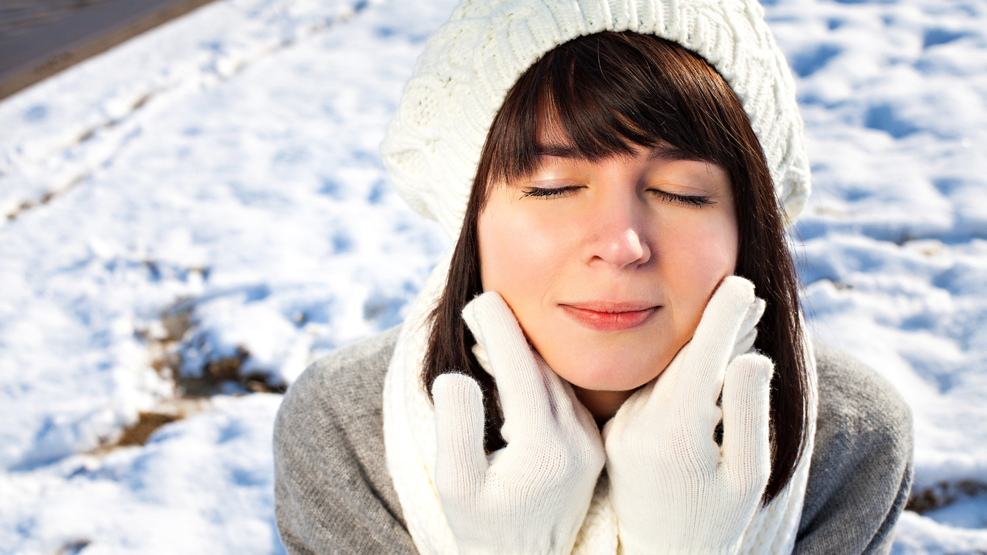 Woman in winter clothing, eyes closed, touching cheeks, snow in background.