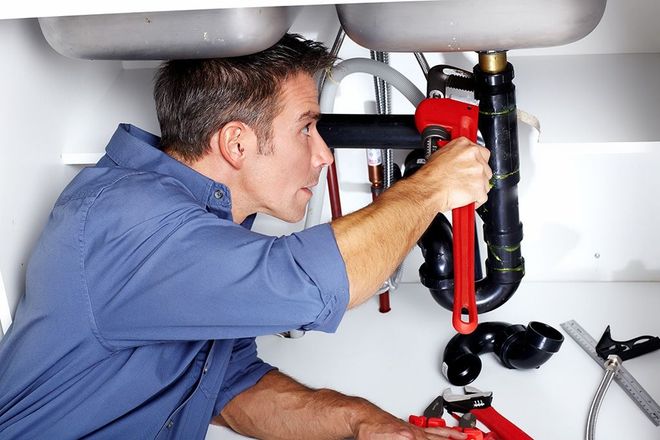 Plumber in blue shirt, using wrench to work under a kitchen sink.