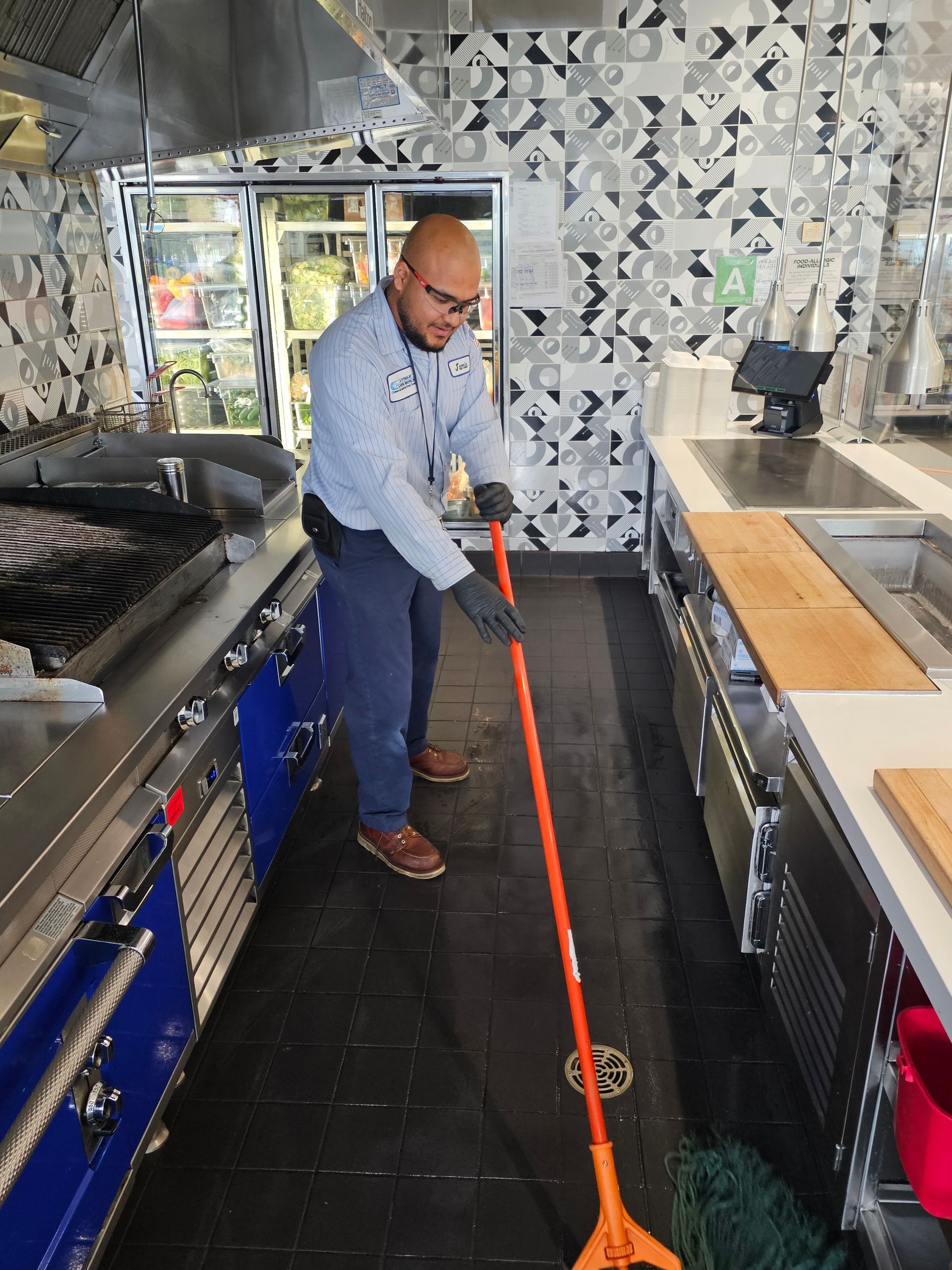Person mopping a restaurant kitchen floor. The kitchen has stainless steel appliances, and blue oven.