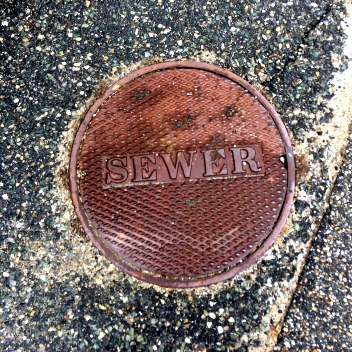 Round, rusty sewer cover embedded in cracked asphalt, reads