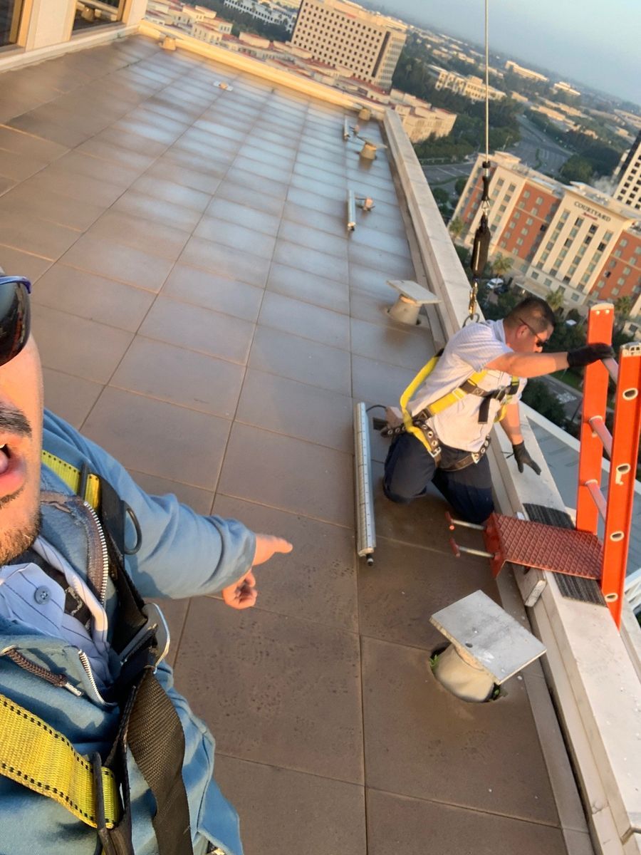 Two people on a rooftop, one pointing, wearing harnesses. City view in background.
