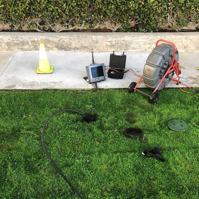 Sewer line inspection equipment on grass: camera, reel, monitor, case, and cone next to sidewalk.