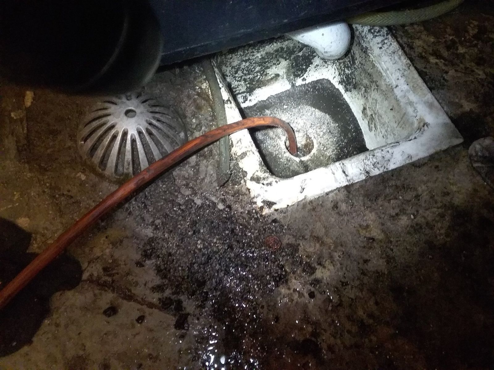 Orange hose draining dirty water into a square floor drain, surrounded by dark debris.