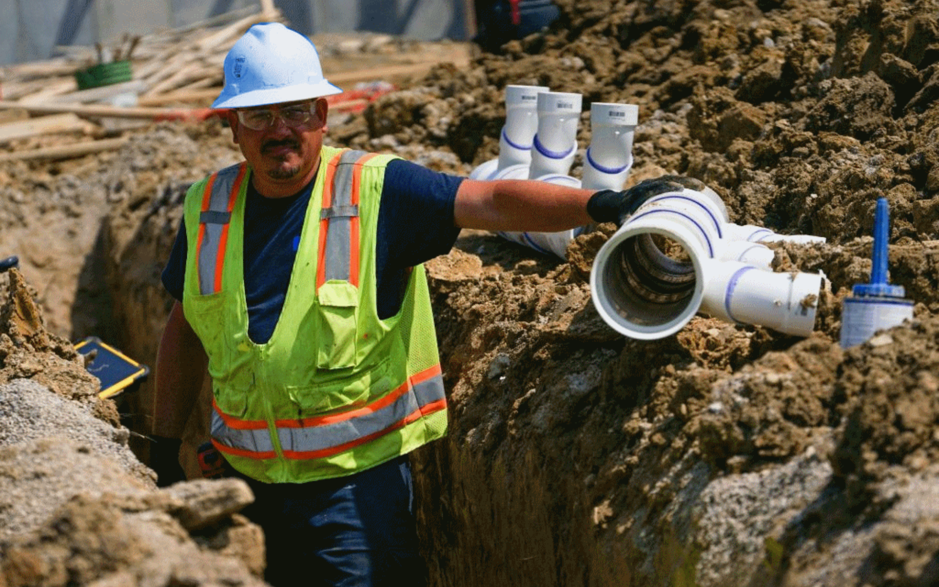 Construction worker in a hard hat and neon vest installing white pipes in a trench