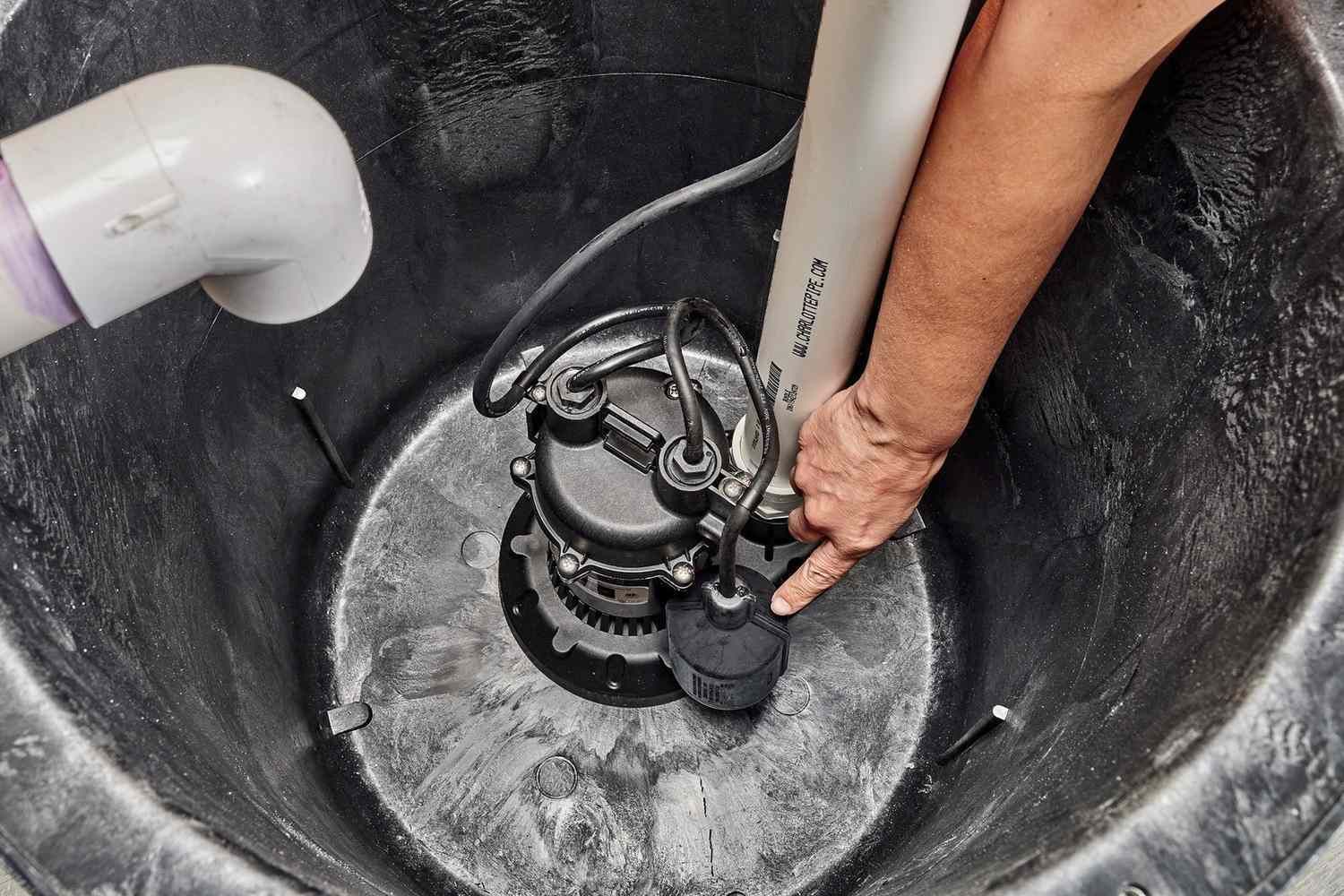 Person installs a sump pump inside a dark, circular basin, with white PVC piping.