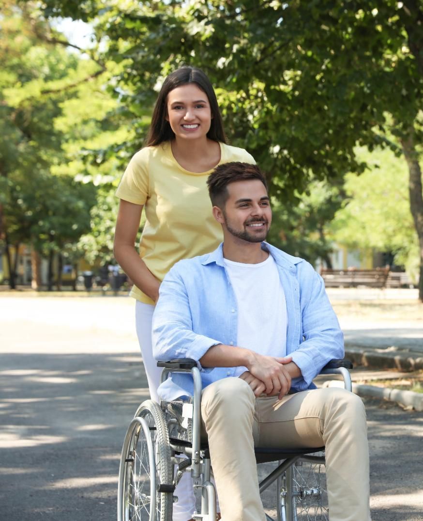 Woman pushing a wheelchair with a man in it along a path, trees in background, sunny day.