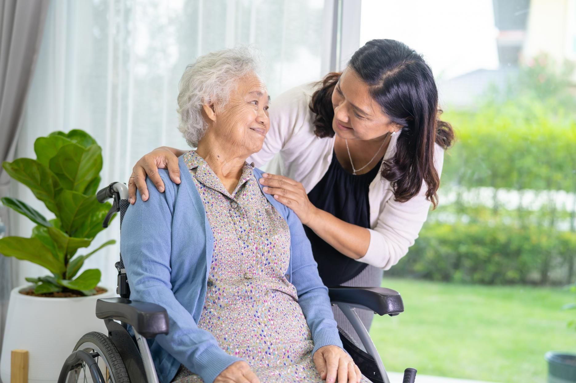 Woman comforts senior in wheelchair, hand on shoulder, looking at each other indoors near a window.