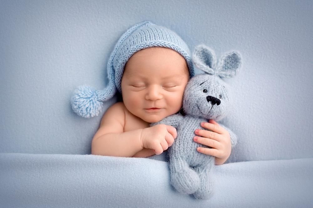Newborn baby wearing blue knitted cap, snuggling blue rabbit toy, sleeping on blue blanket.