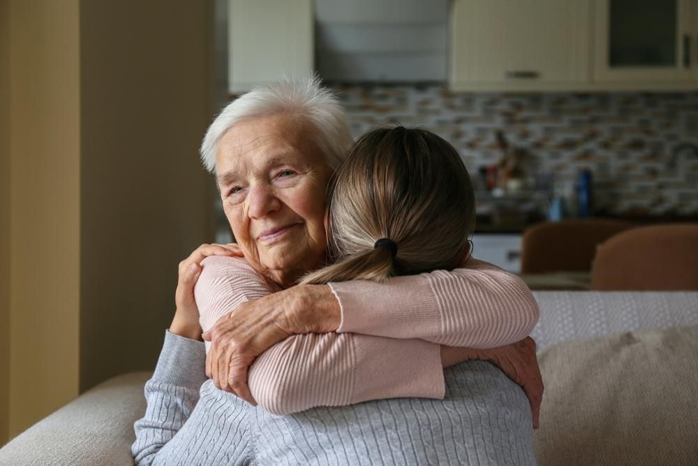 An older person and a younger person embrace on a sofa in a home setting.