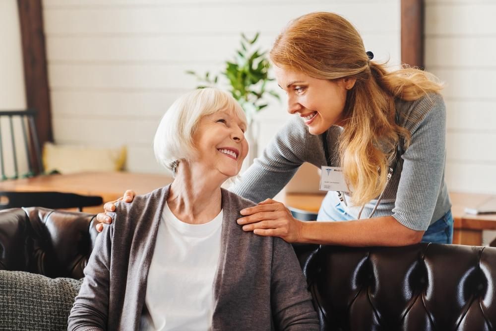 Woman comforts older person on couch, both smiling. The woman has an arm around the other.